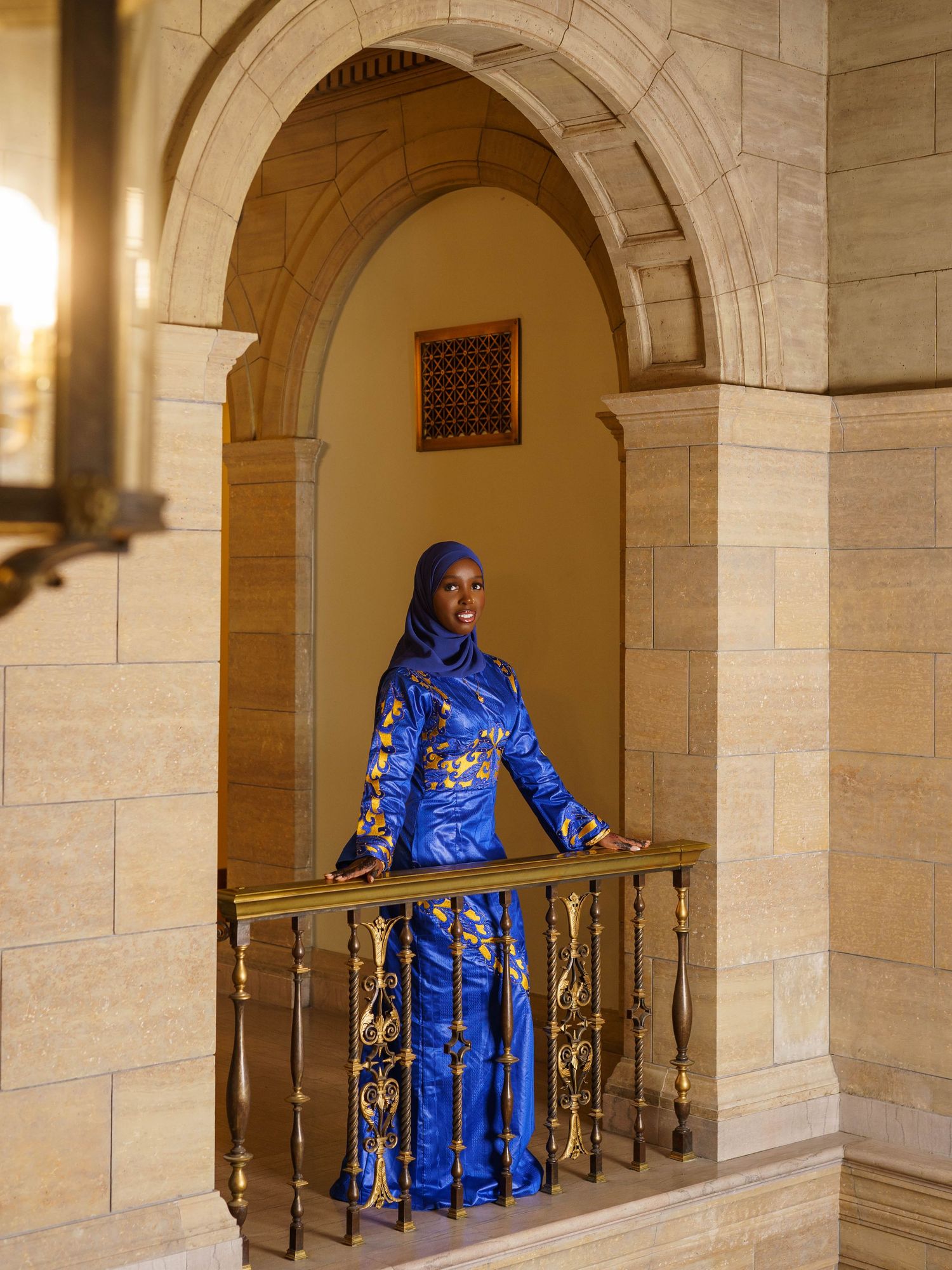 Person wearing bright blue traditional dress standing in an ornate stone archway with decorative railing.
