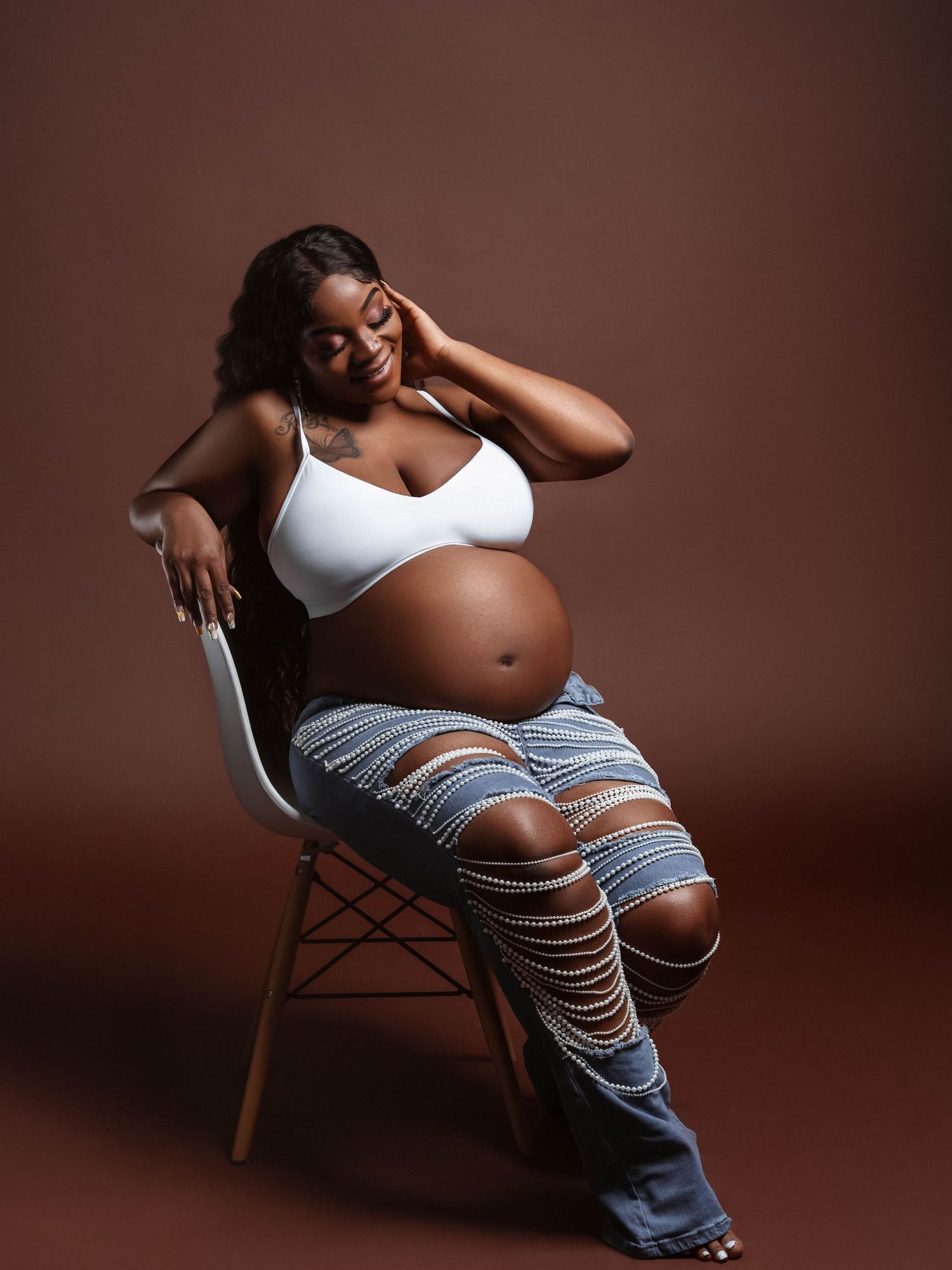 Artistic maternity portrait with pearl strands and distressed denim against brown backdrop.