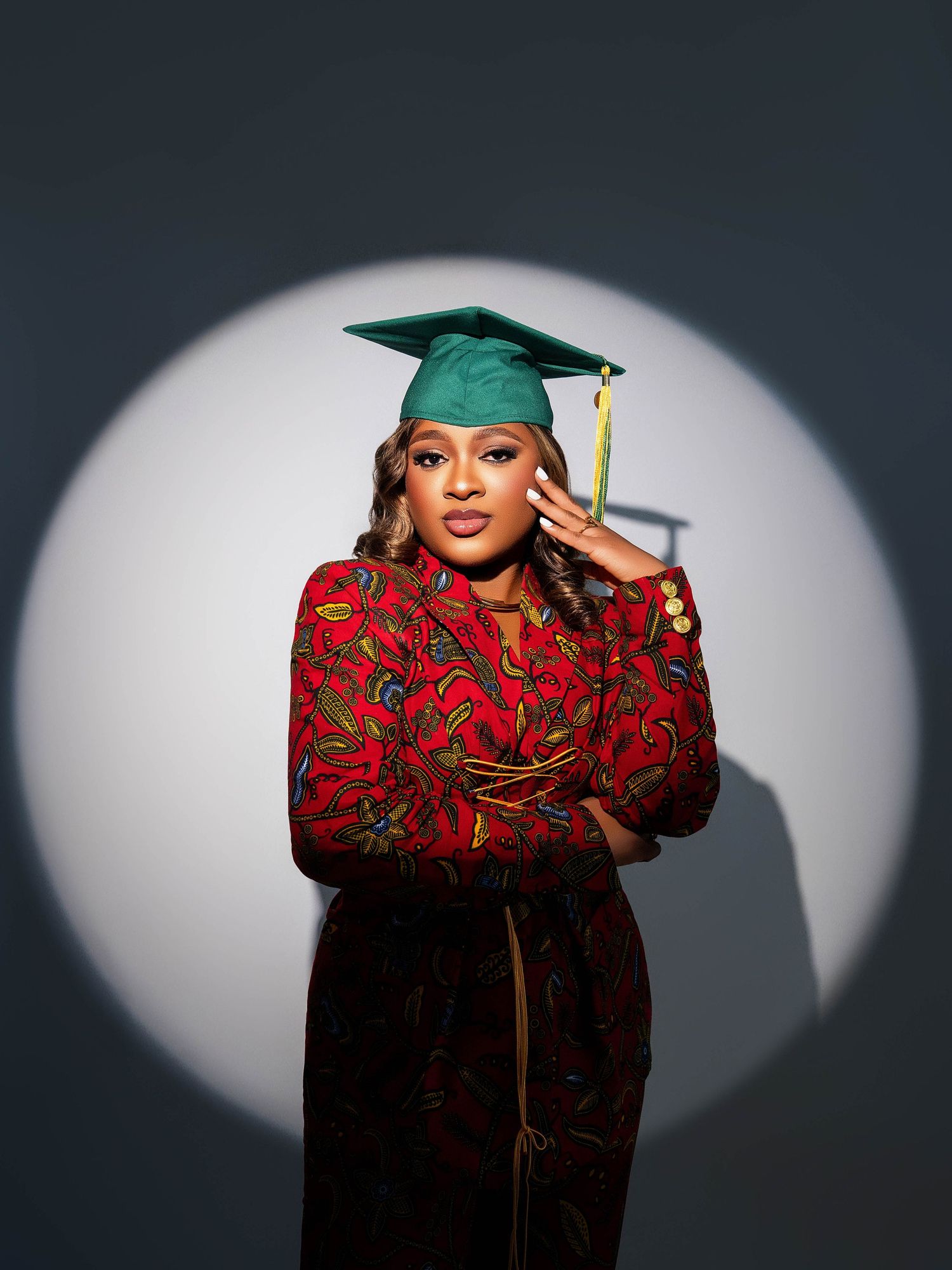 Graduation portrait featuring graduate in teal cap and red floral dress against dramatic spotlight backdrop.