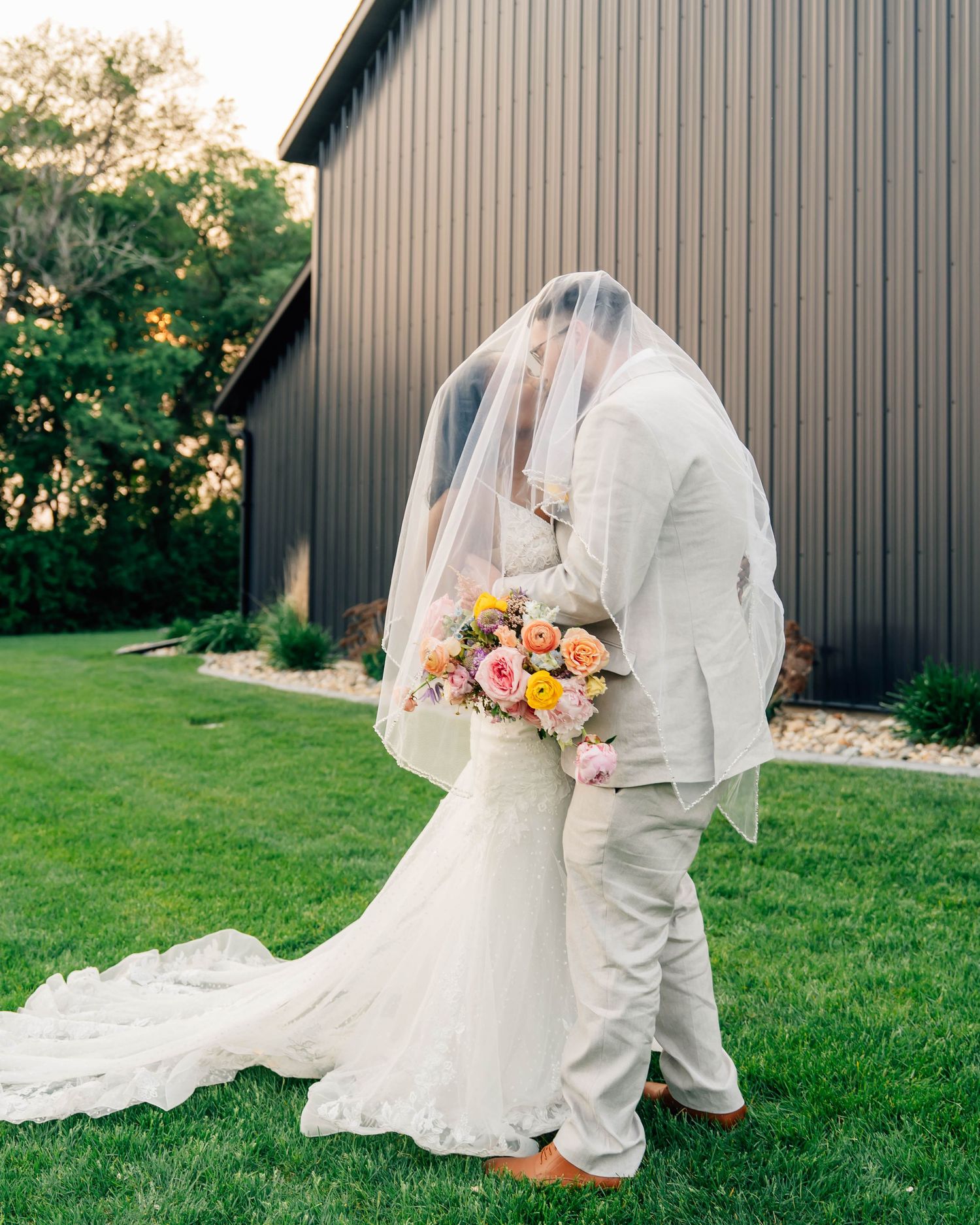 Wedding couple poses against modern barn backdrop with bride's flowing white dress.