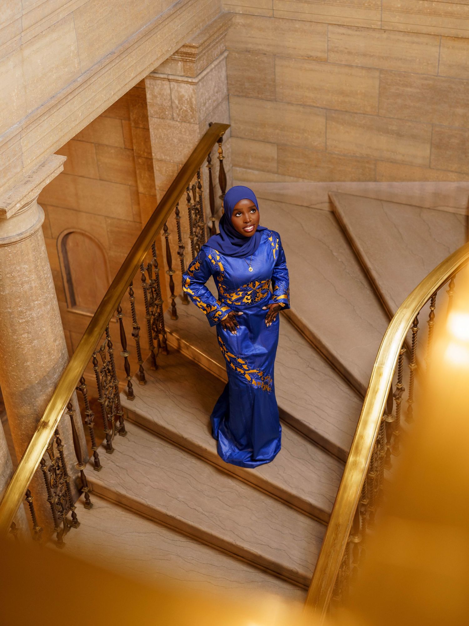 Elegant pose on grand marble staircase wearing royal blue embroidered dress with hijab.