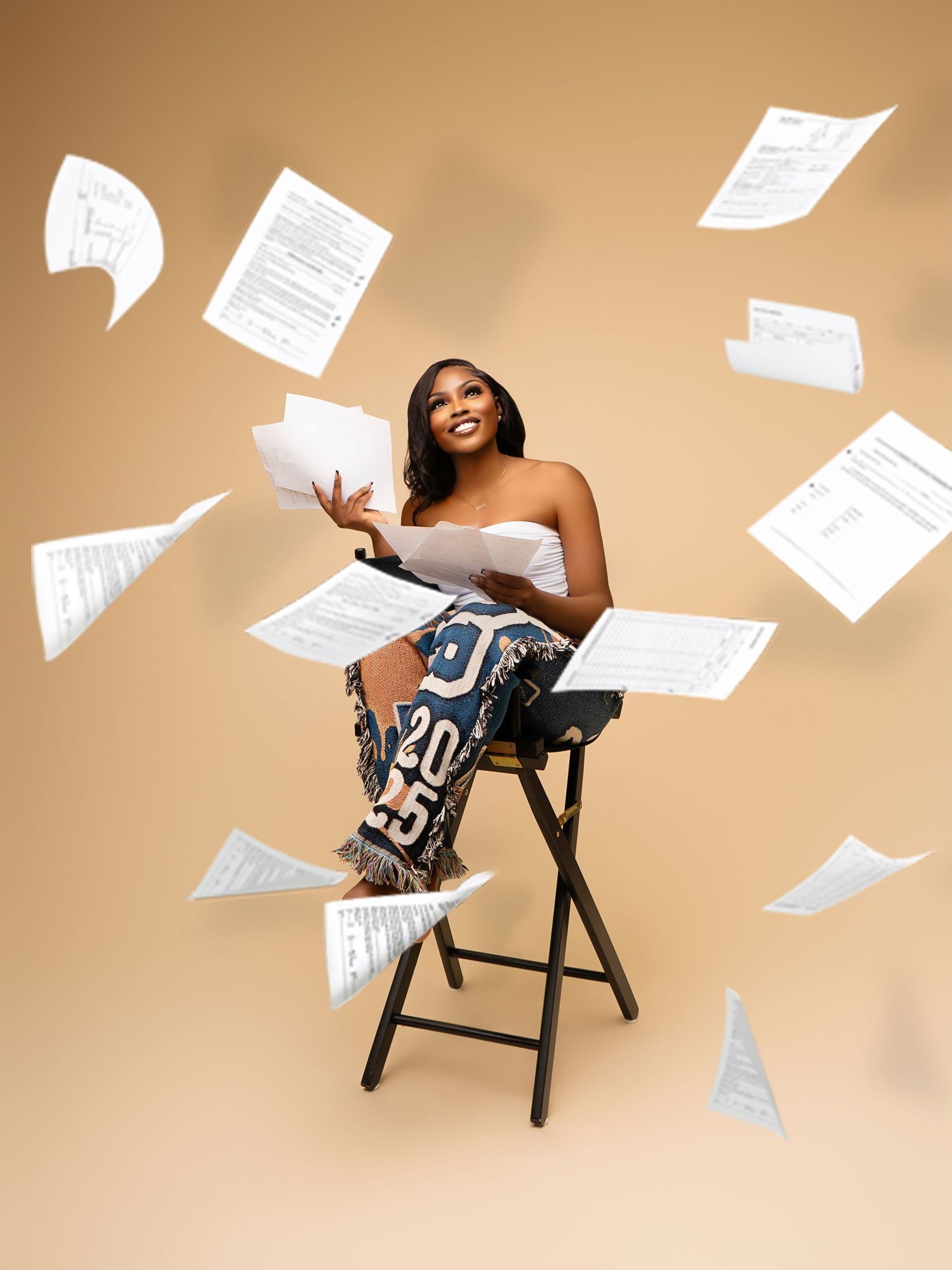 Creative portrait with floating papers around woman sitting on stool against beige background.