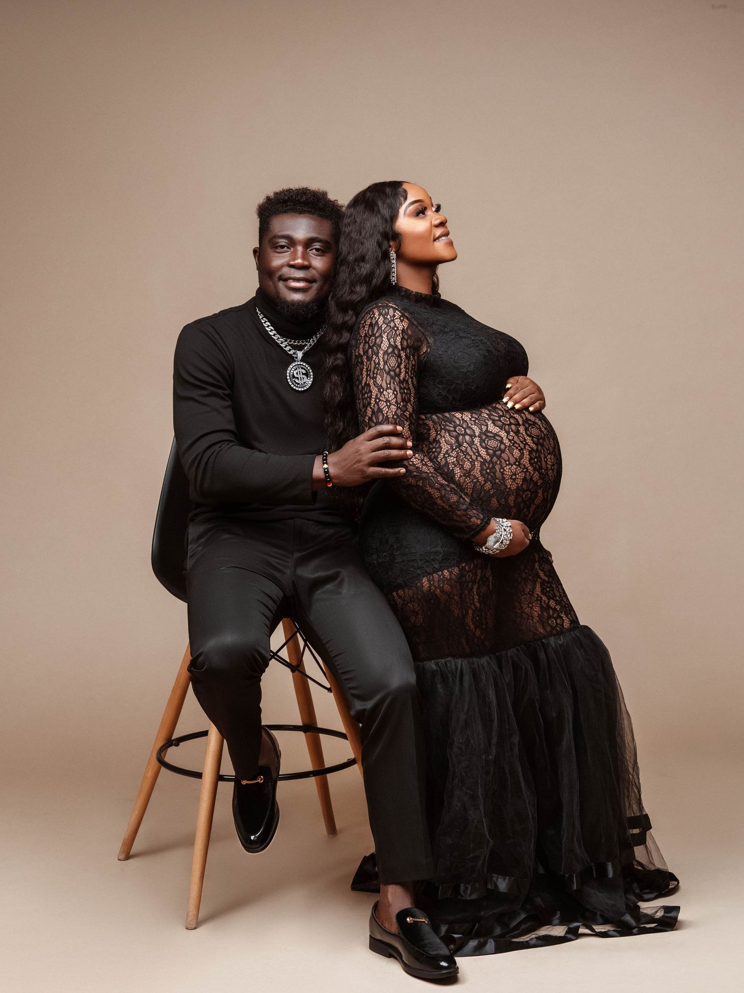 Formal couple in black attire pose intimately on wooden chairs against neutral backdrop.