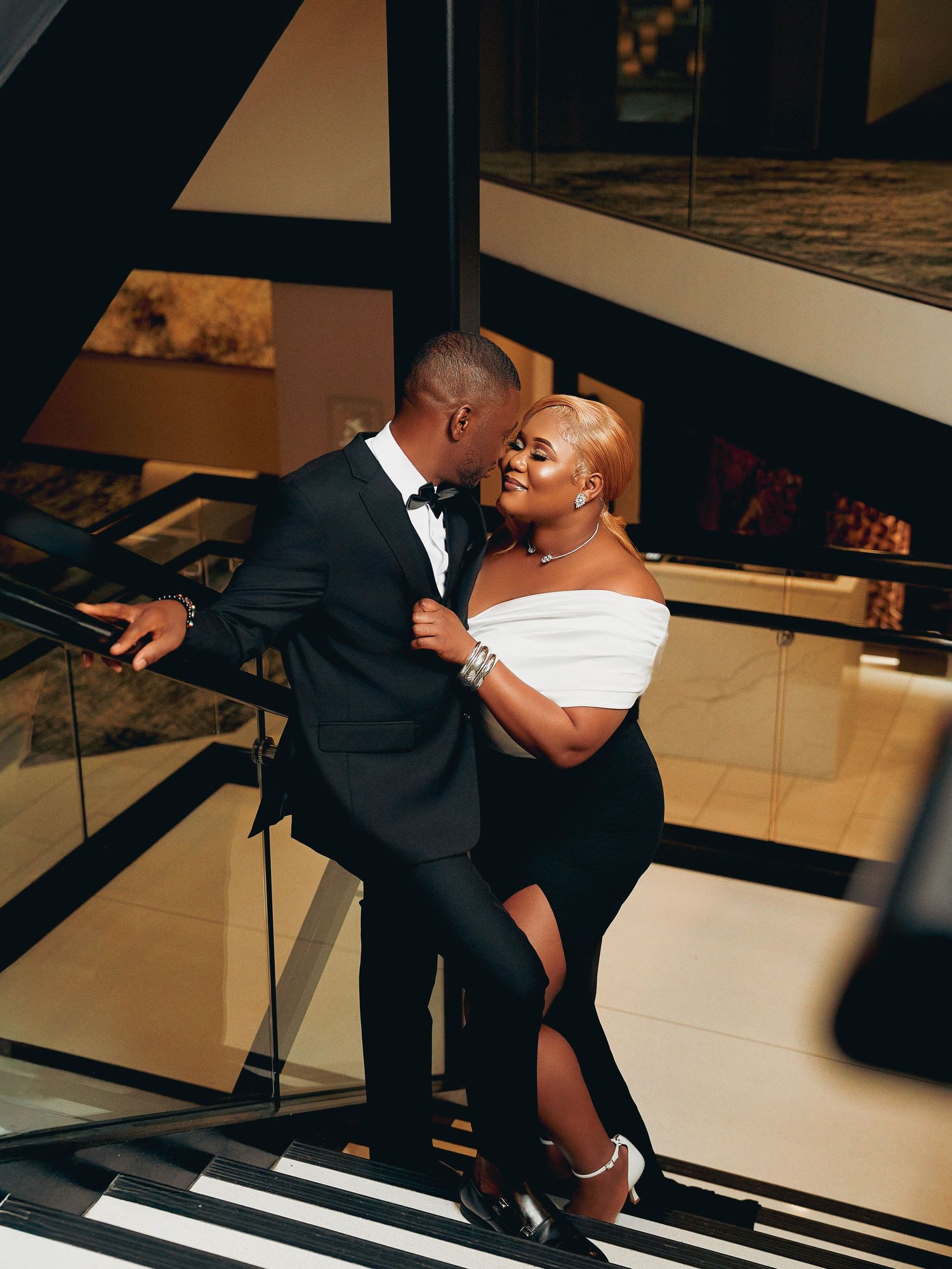 Elegant couple sharing intimate moment on modern staircase in formal attire.