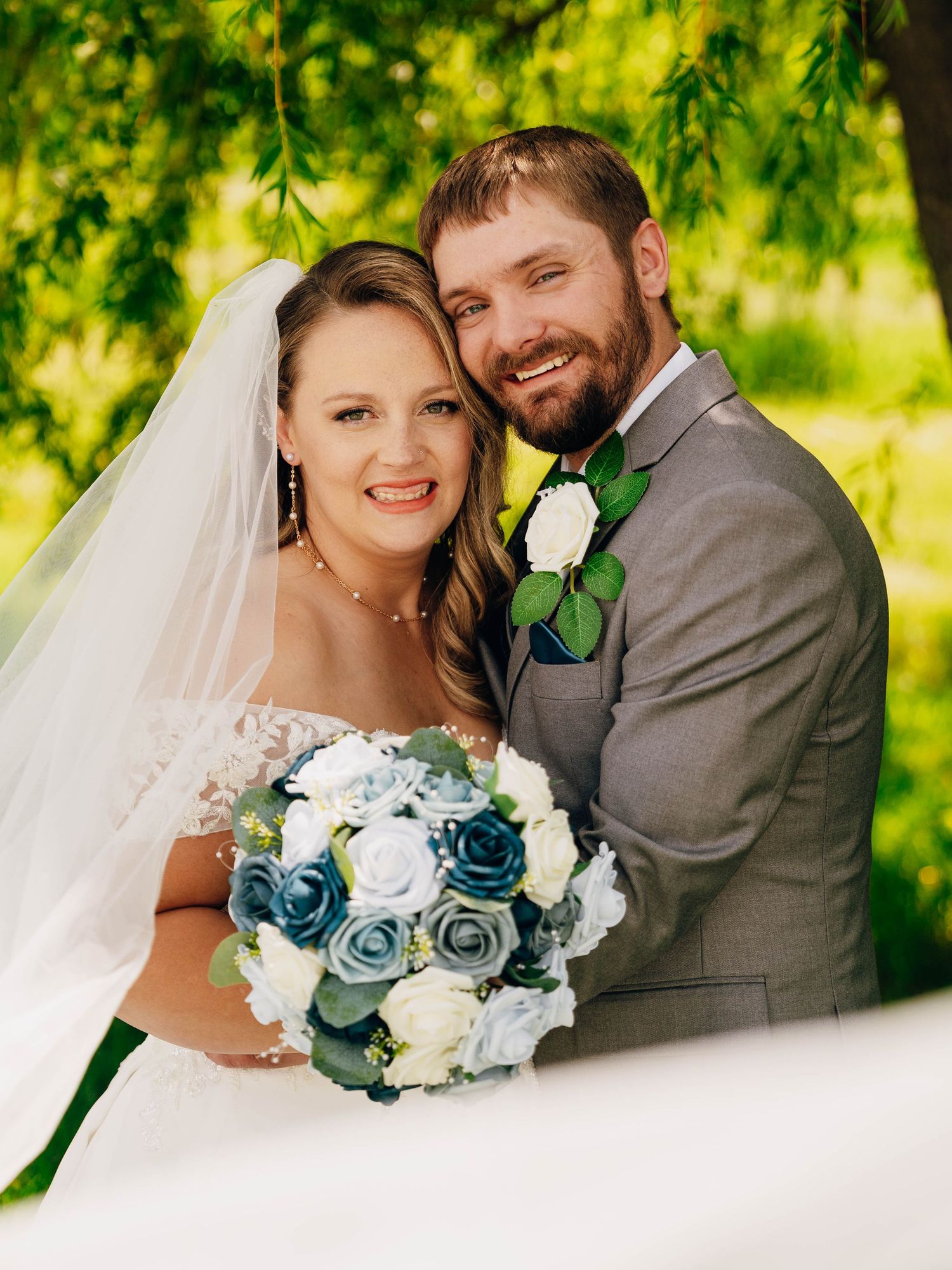 Wedding couple in grey suit and white dress pose with blue and white bouquet under green willow tree branches.