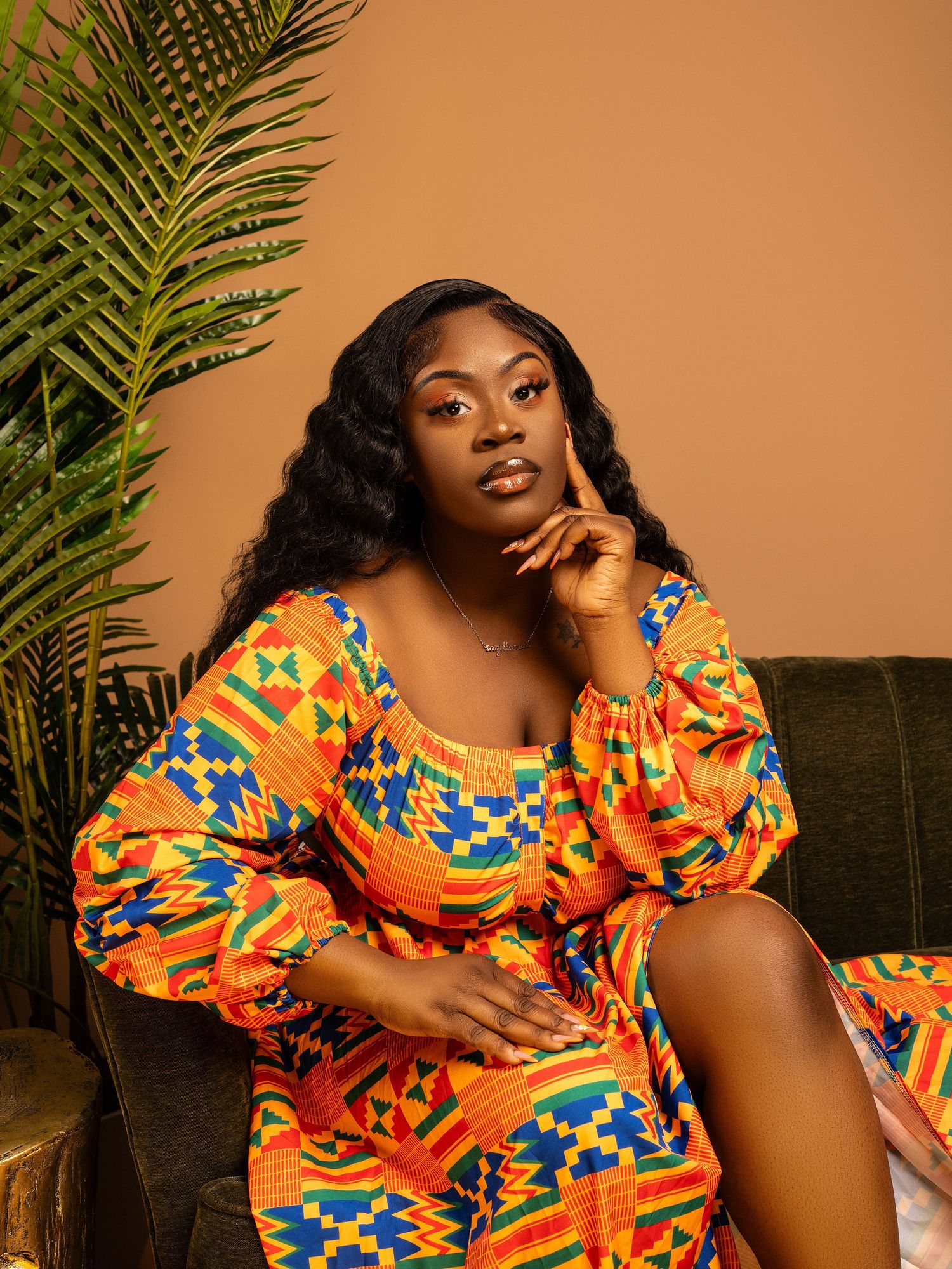 Person wearing colorful geometric patterned dress lounges on dark couch with palm fronds in background.