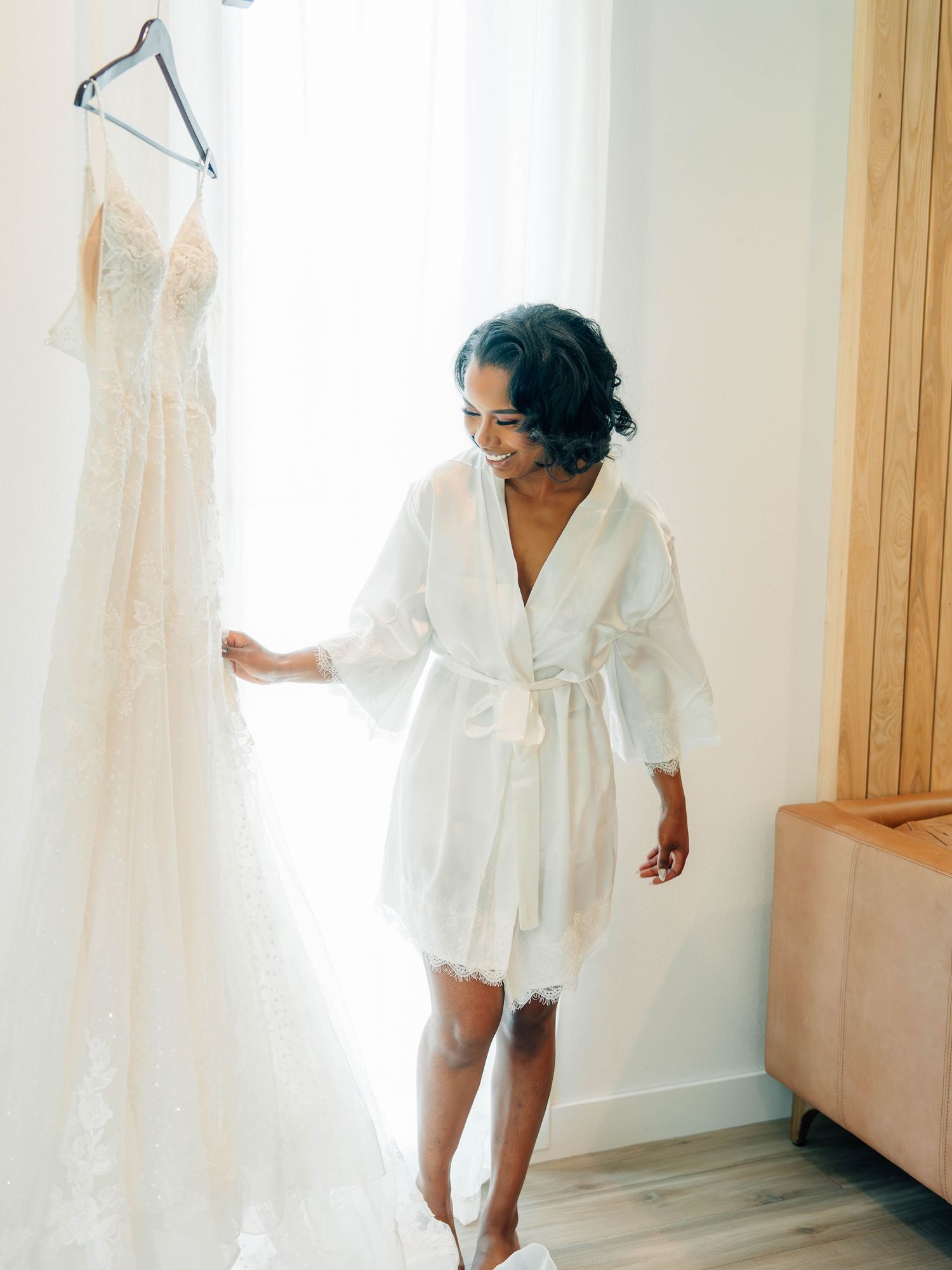 Bride in white silk robe admires wedding dress hanging on display in bridal suite.