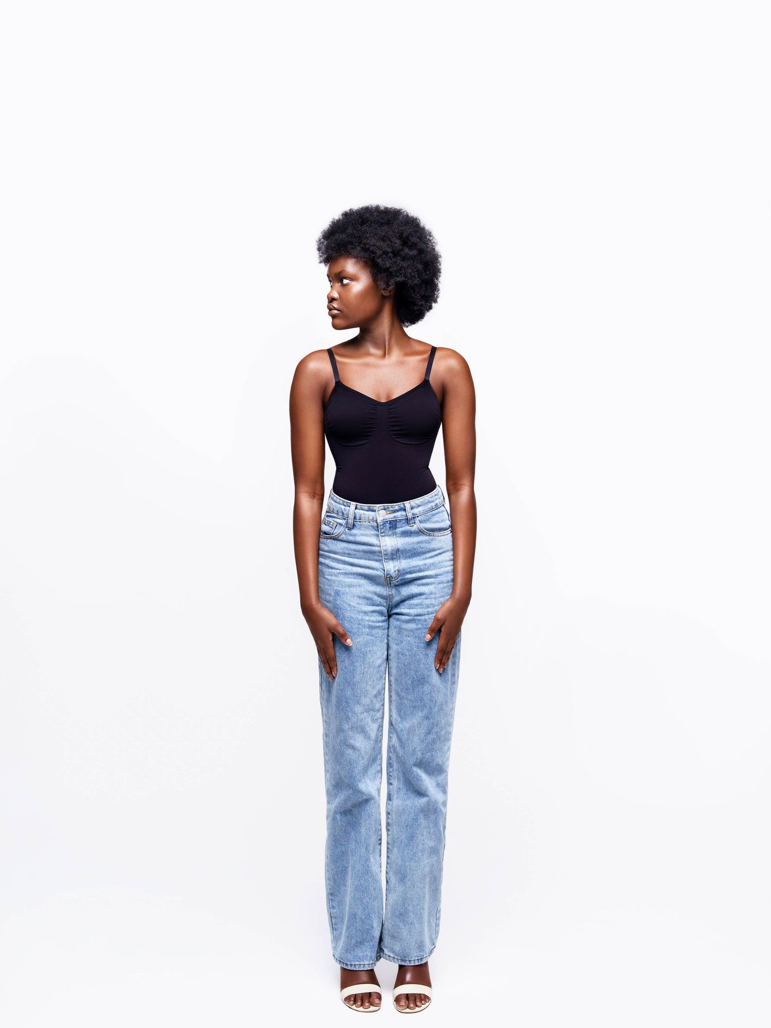 Fashion model poses in black tank top and light wash jeans against white background.