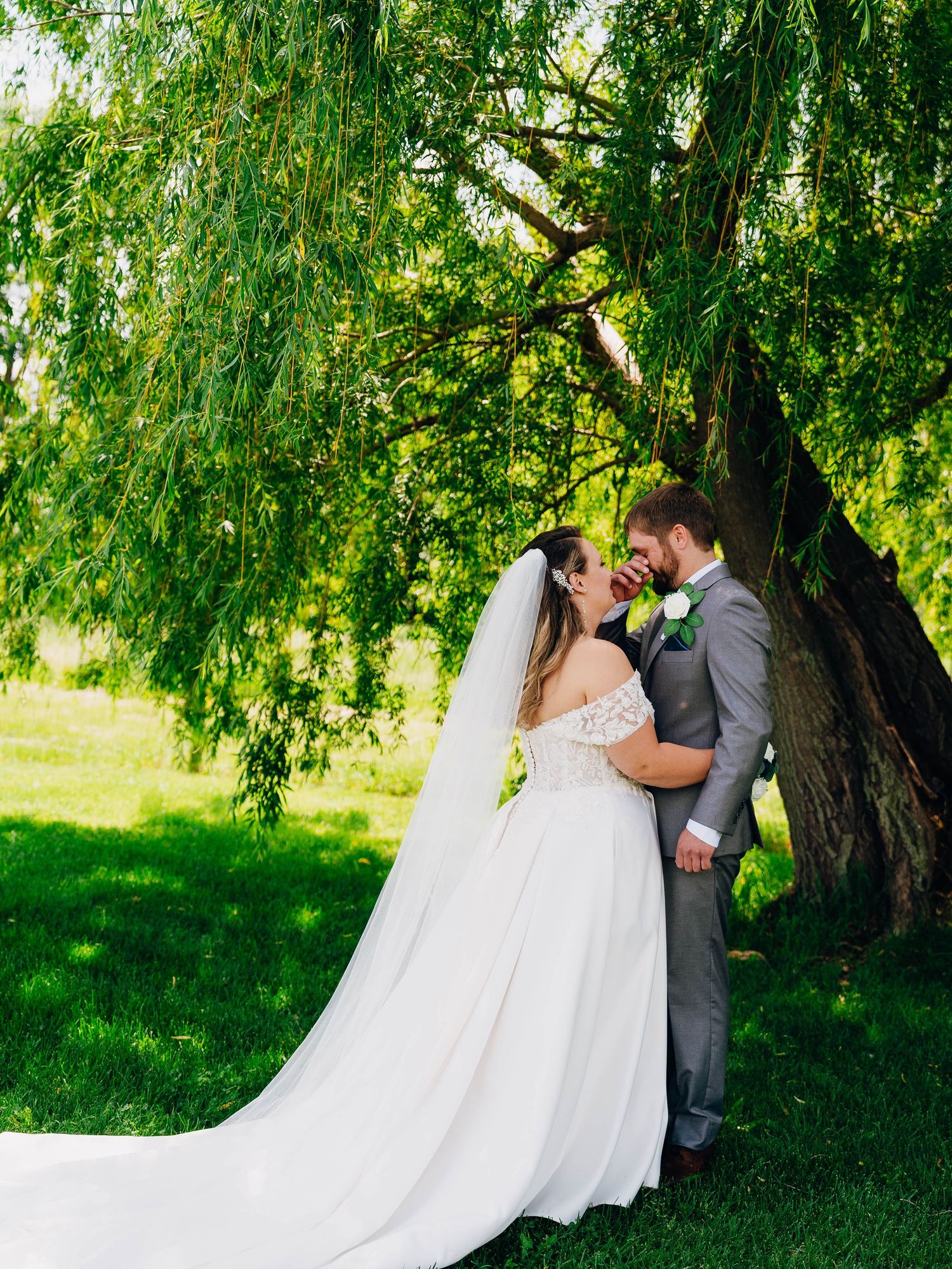 A romantic wedding couple embraces under a sweeping willow tree on a lush green lawn.