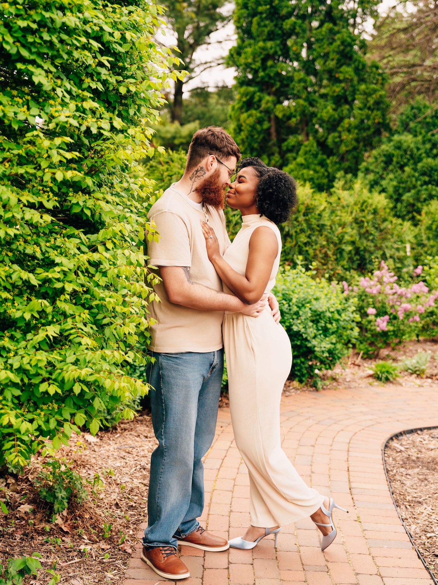 A romantic couple embraces on a garden path surrounded by lush green foliage and flowering plants.
