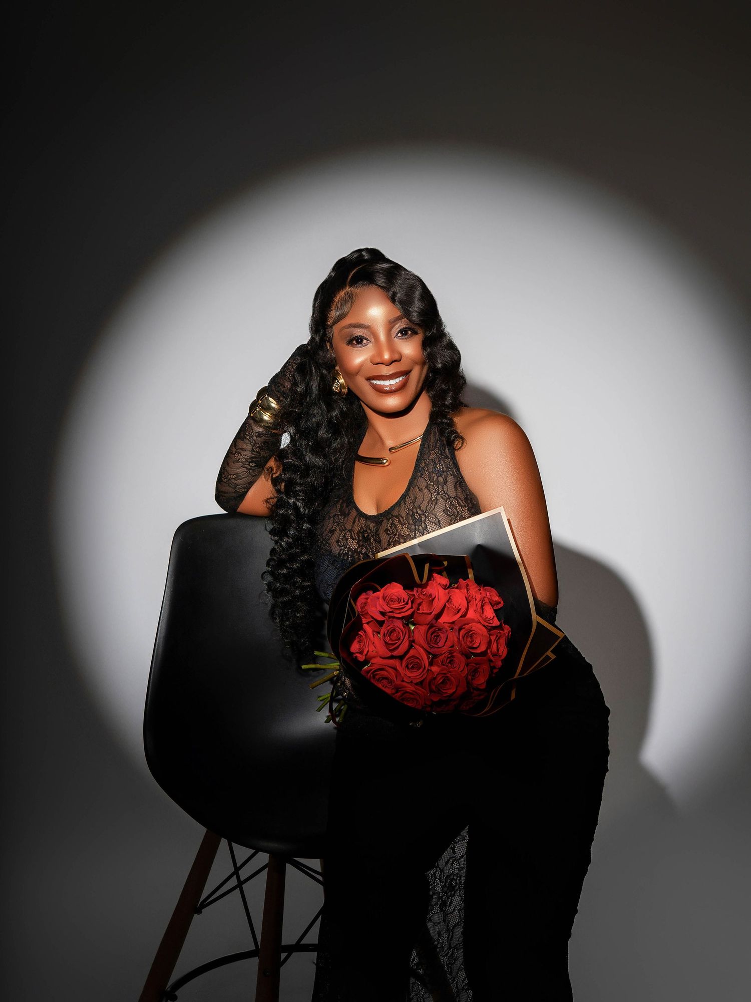 Spotlight portrait with black dress holding red roses on dark chair against white background.
