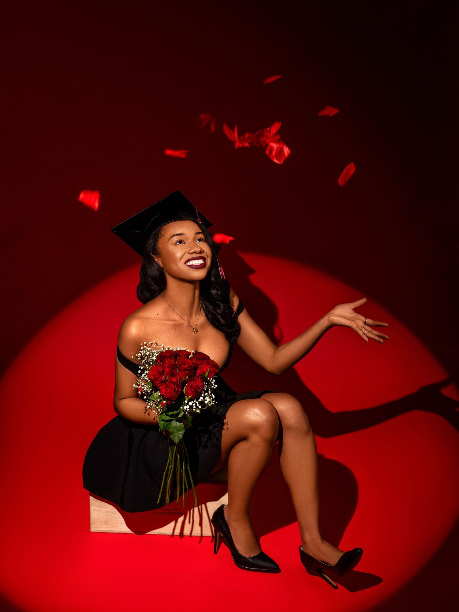 A graduation celebration photo with red roses and cap against a dramatic red spotlight background.