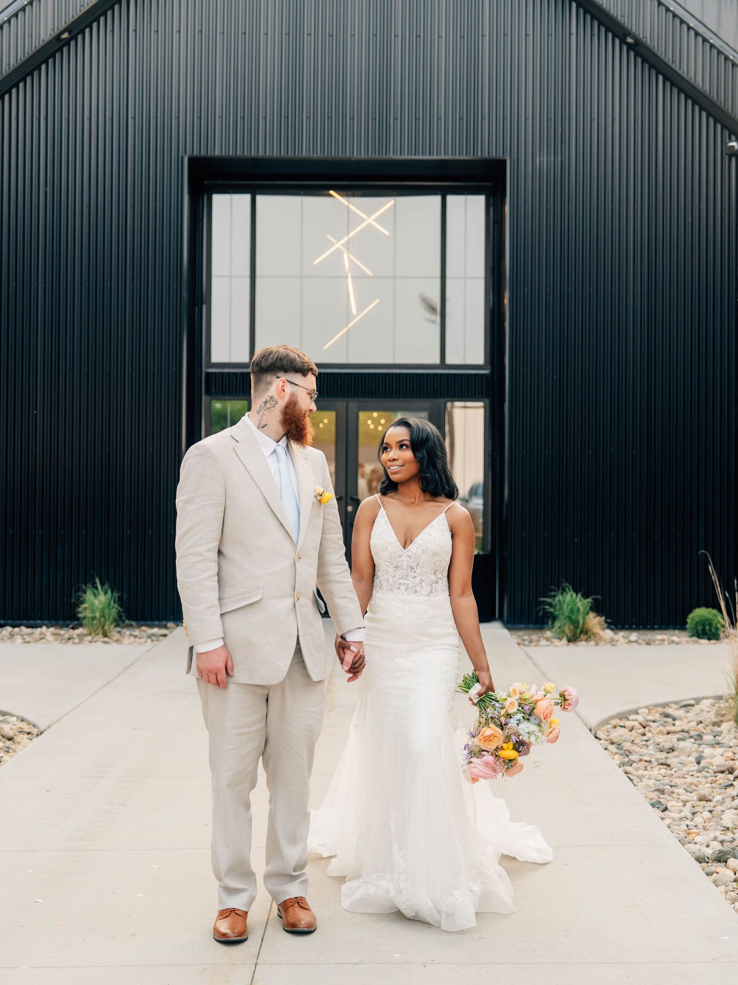 Modern wedding portraits in front of industrial black building with white dress and light suit.