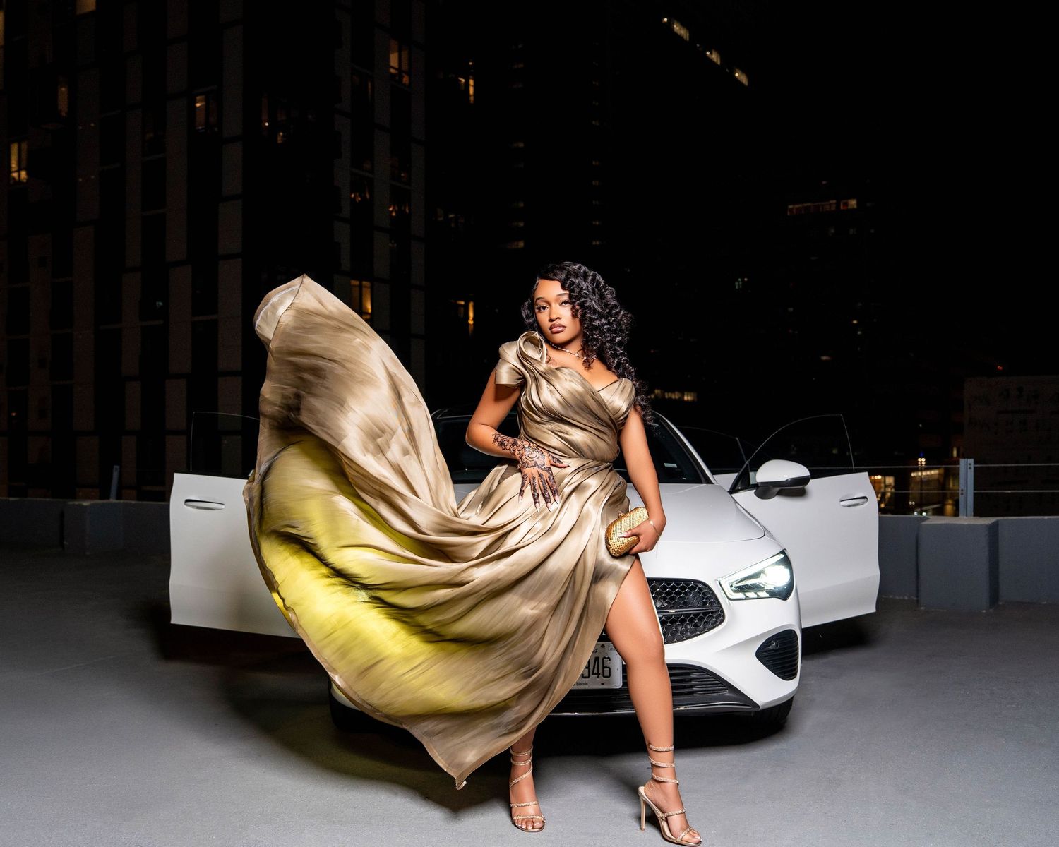 Glamorous fashion shot with flowing gold dress and white luxury car in dark urban setting.