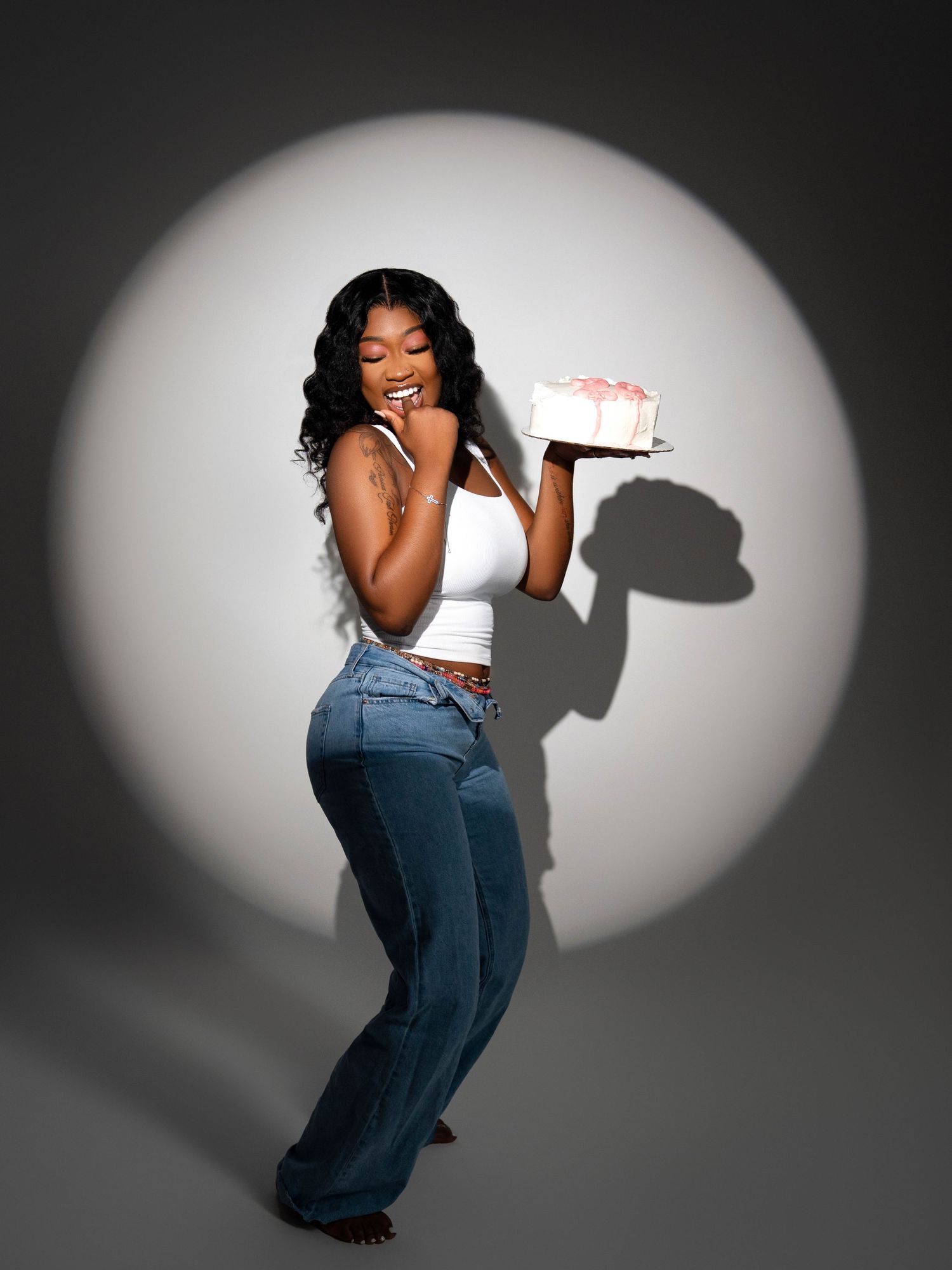 Joyful portrait of person in casual outfit holding small cake against white circular spotlight background.
