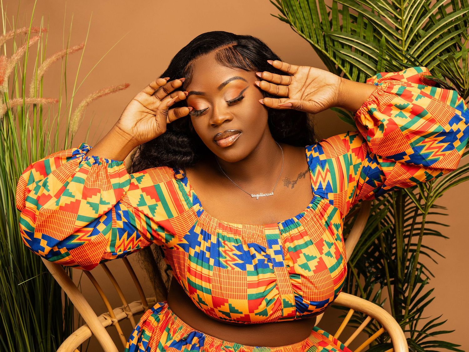 Model in vibrant orange African print dress poses gracefully against warm backdrop with tropical plants.