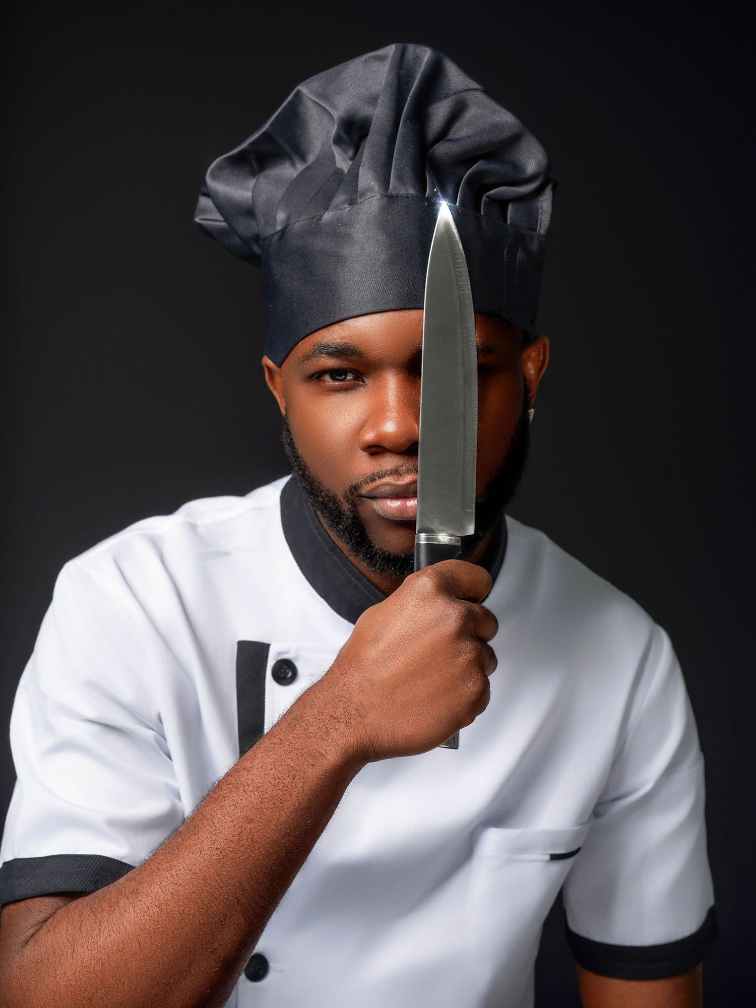 Professional chef in white uniform and black hat poses with knife in dramatic studio lighting.