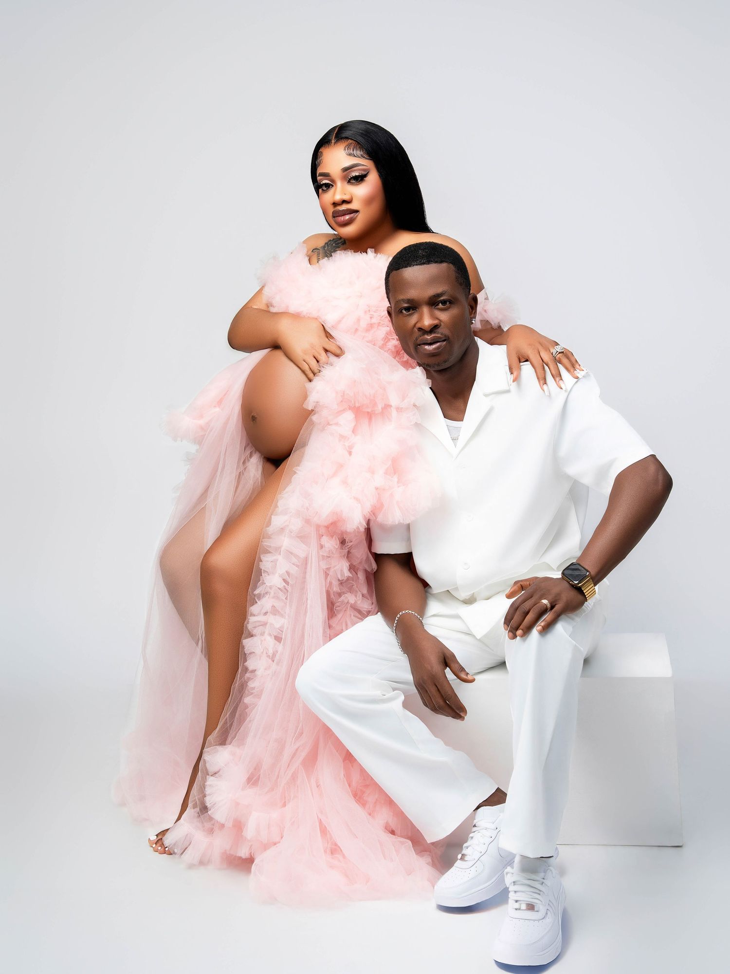 A romantic couple poses in a studio portrait wearing coordinated white and pink outfits against a light backdrop.