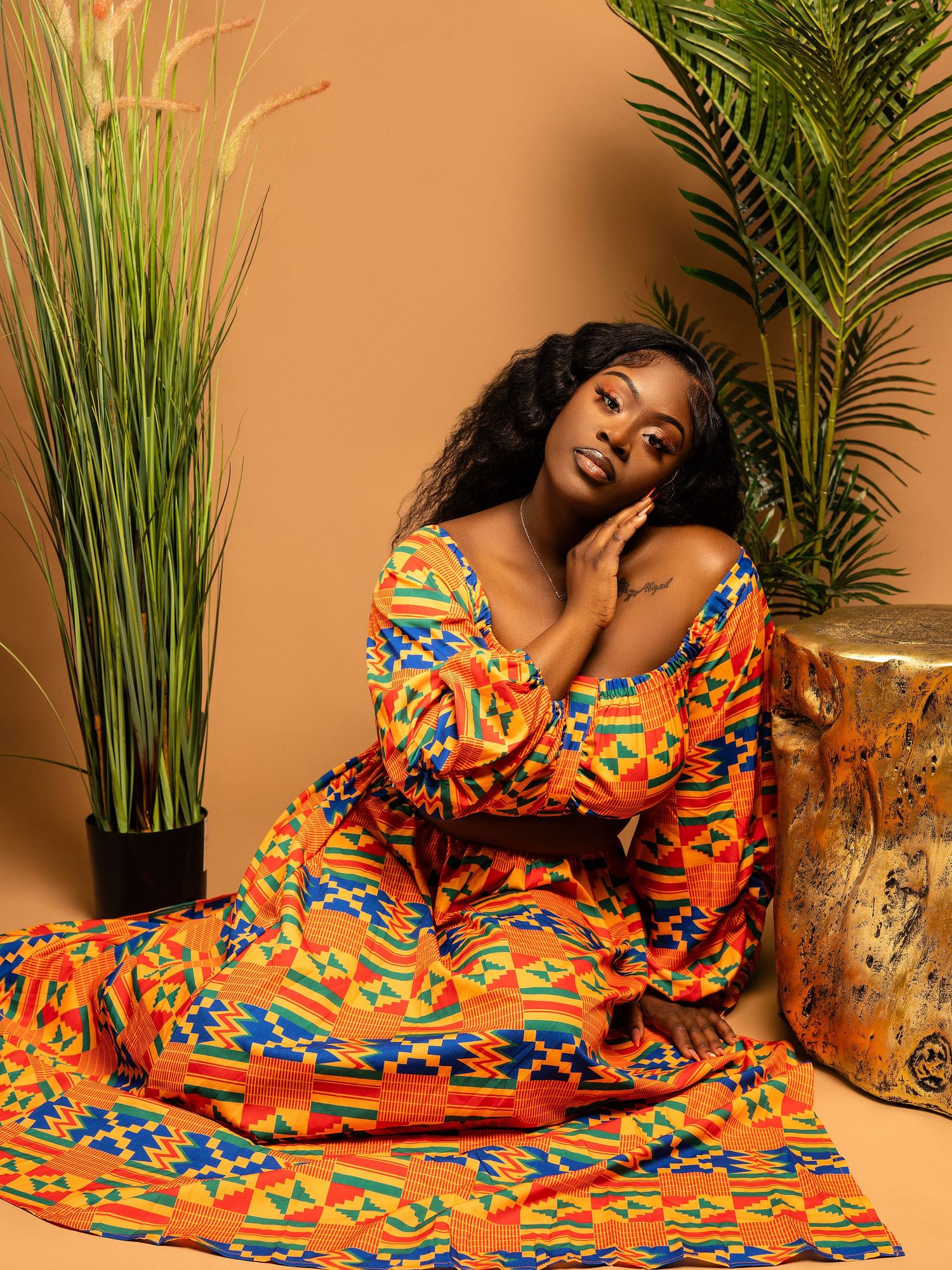 Model in bold orange African print dress reclines elegantly on sofa with decorative palm plants.