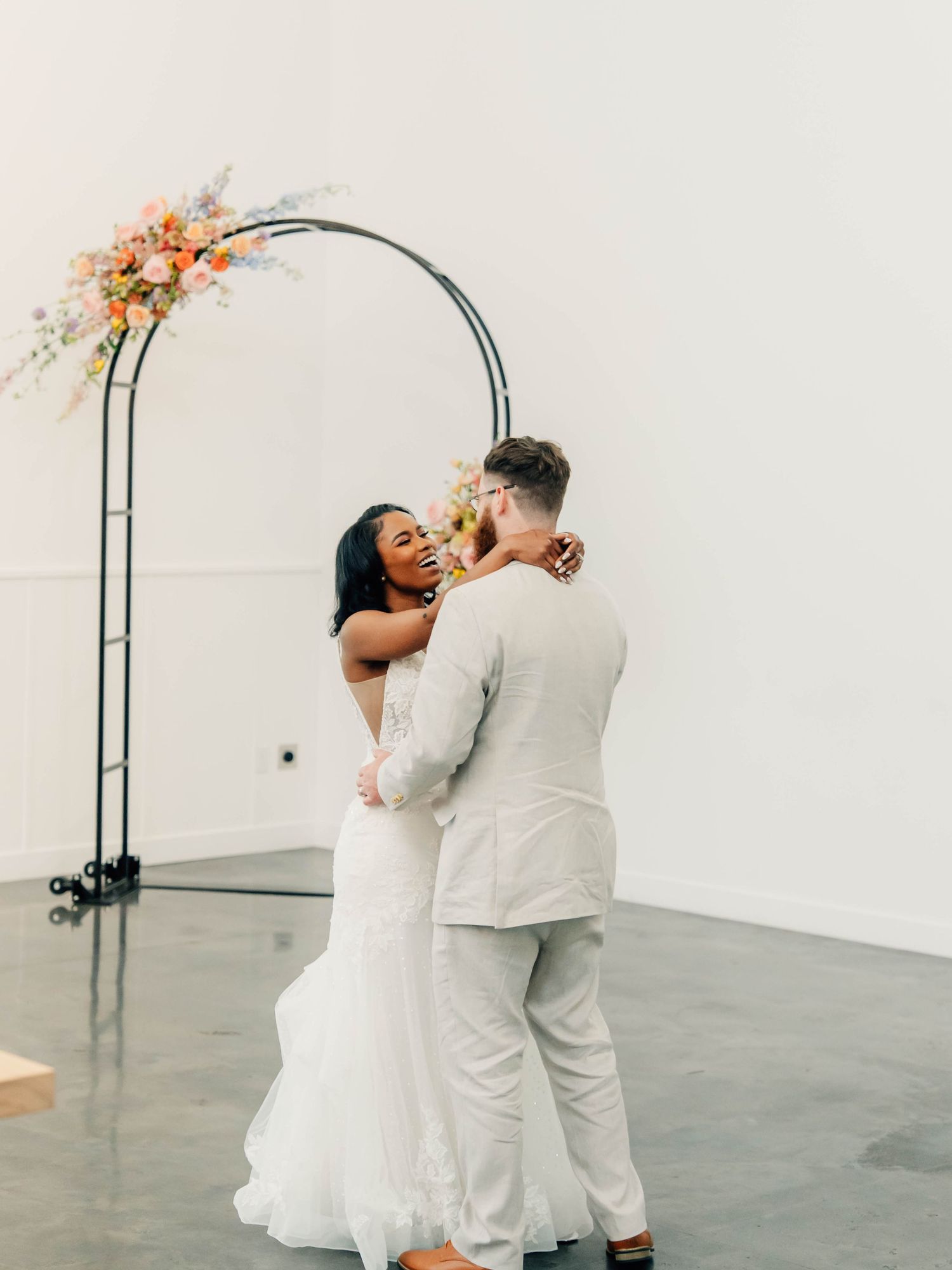 Wedding ceremony under floral arch with coral and peach blooms against white wall.