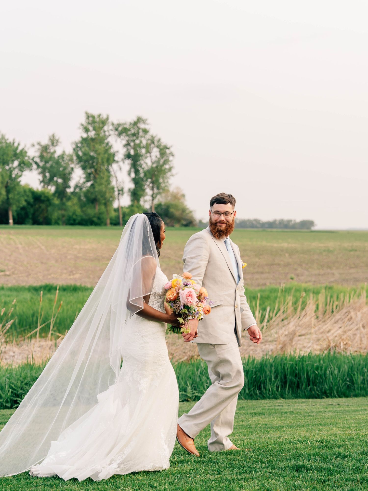 Wedding couple walking through rustic outdoor field with flowing veil and peach bouquet.