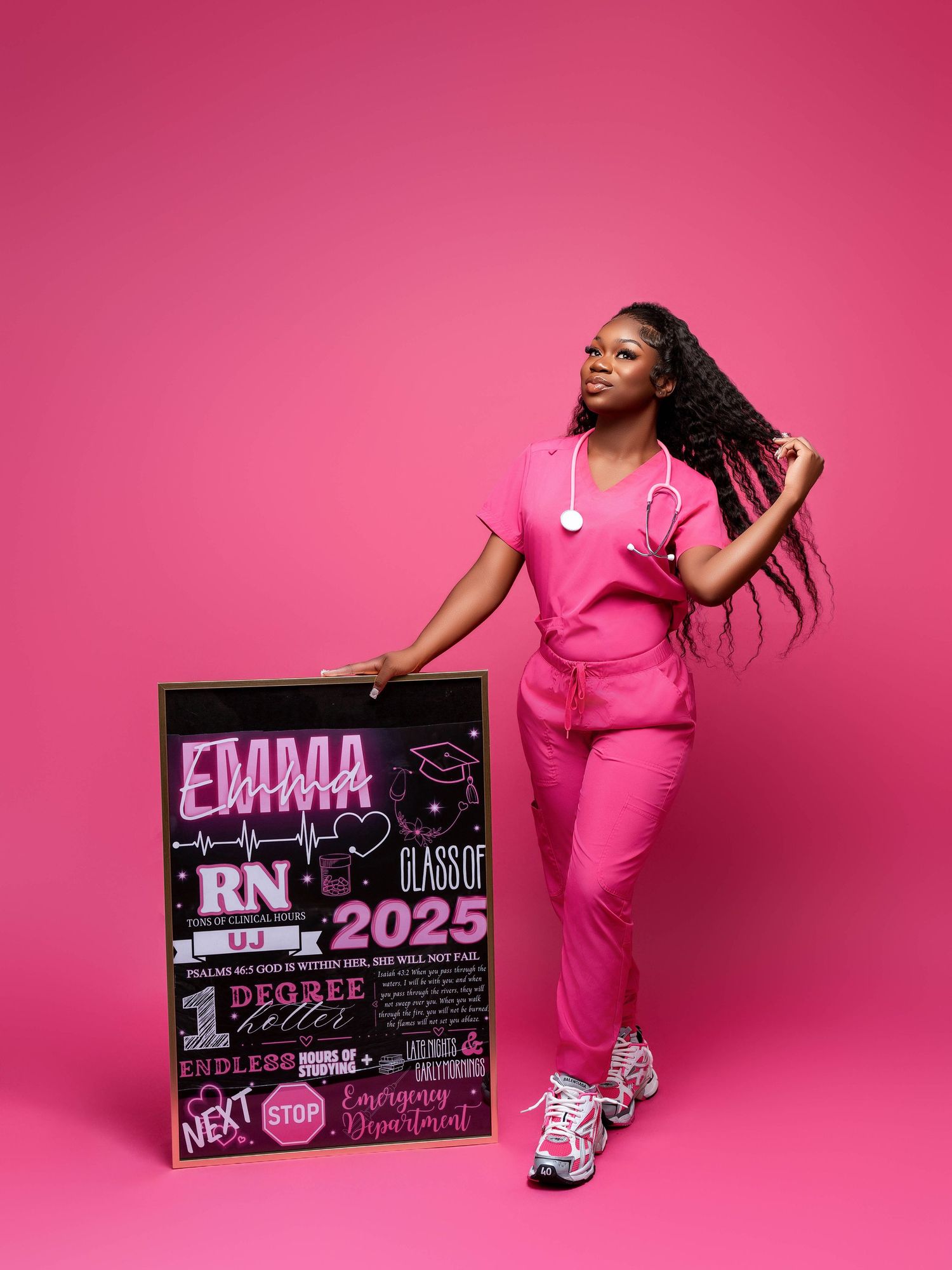 Nurse in pink uniform poses with graduation announcement board showing RN class of 2025.