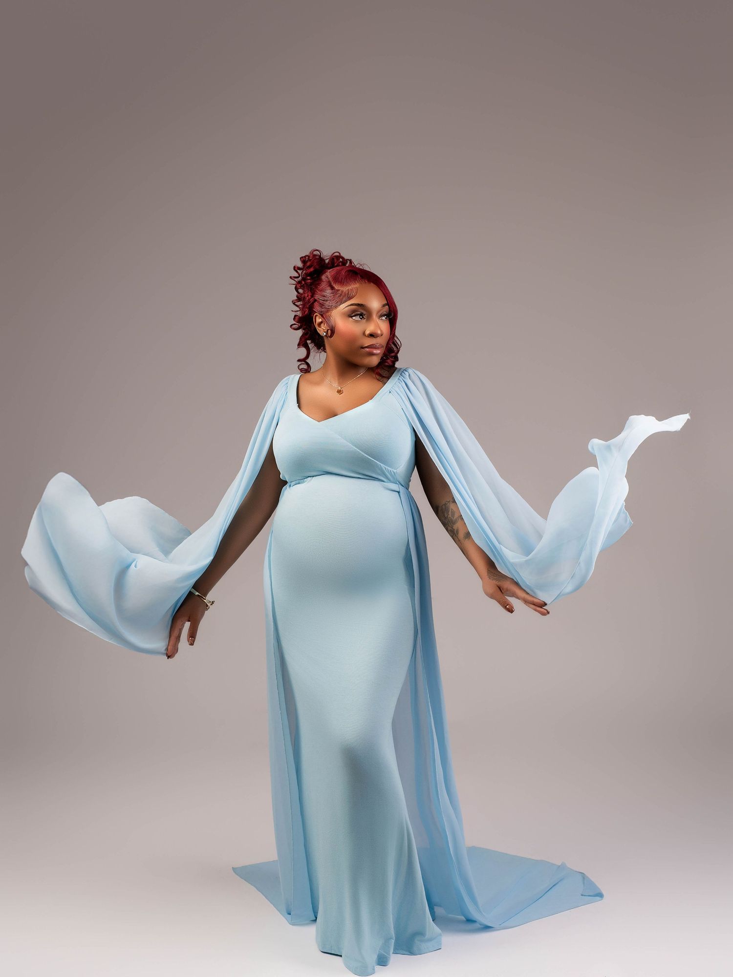 Maternity photo showing flowing light blue dress with dramatic sleeves against grey background.