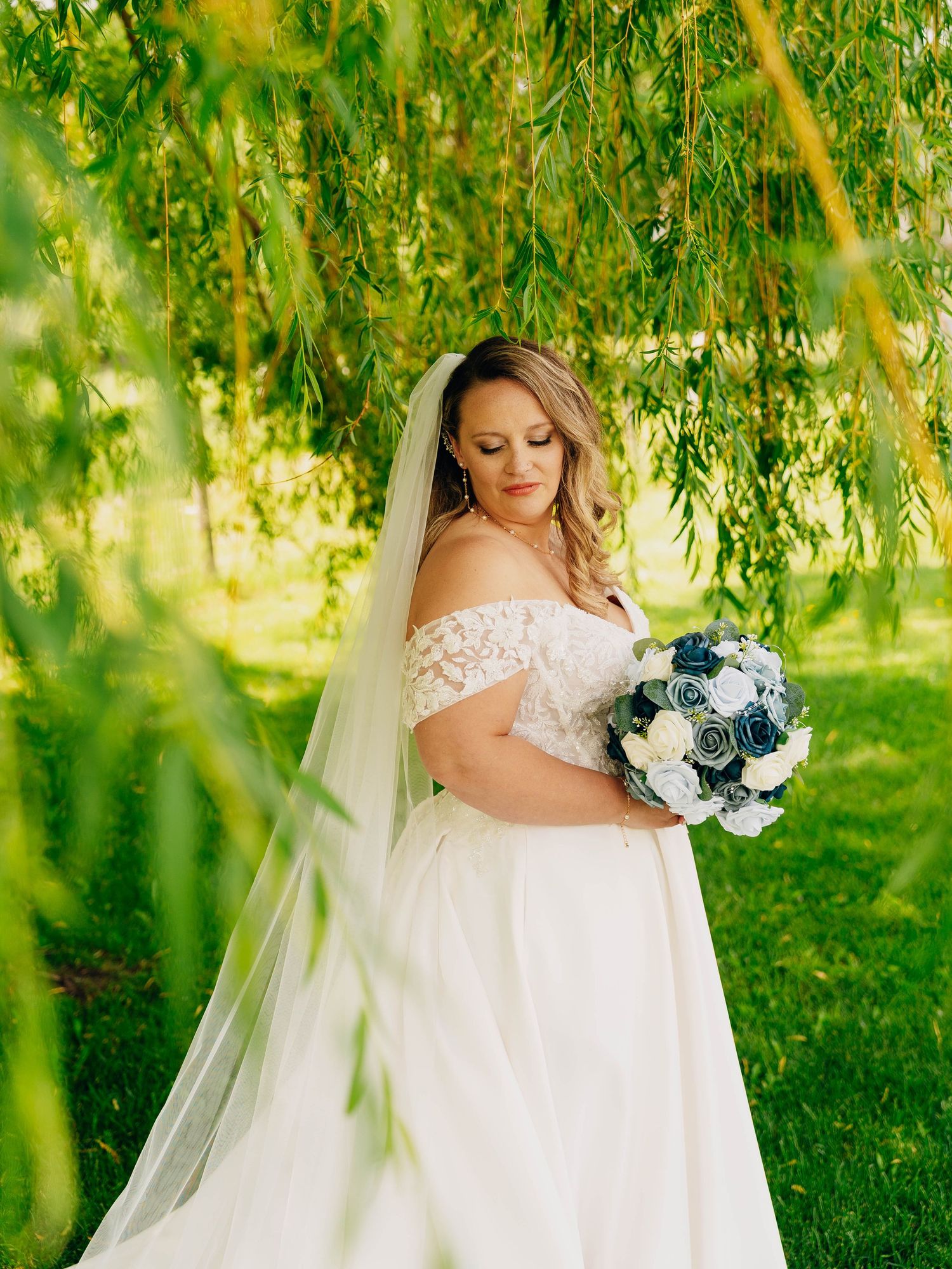 Bride in off-shoulder wedding gown holding bouquet under weeping willow branches.
