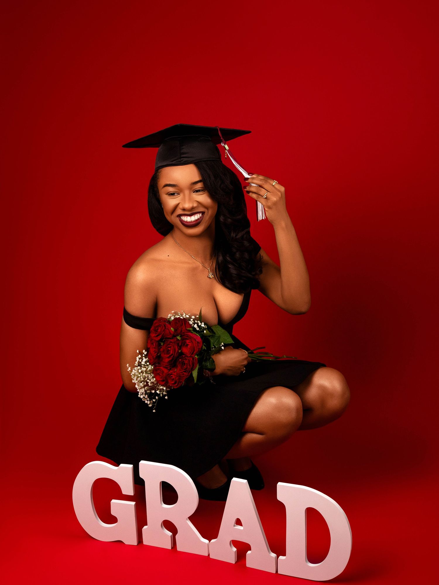 Graduation photo featuring a black dress, cap and gown, red roses and white GRAD letters against a red backdrop.