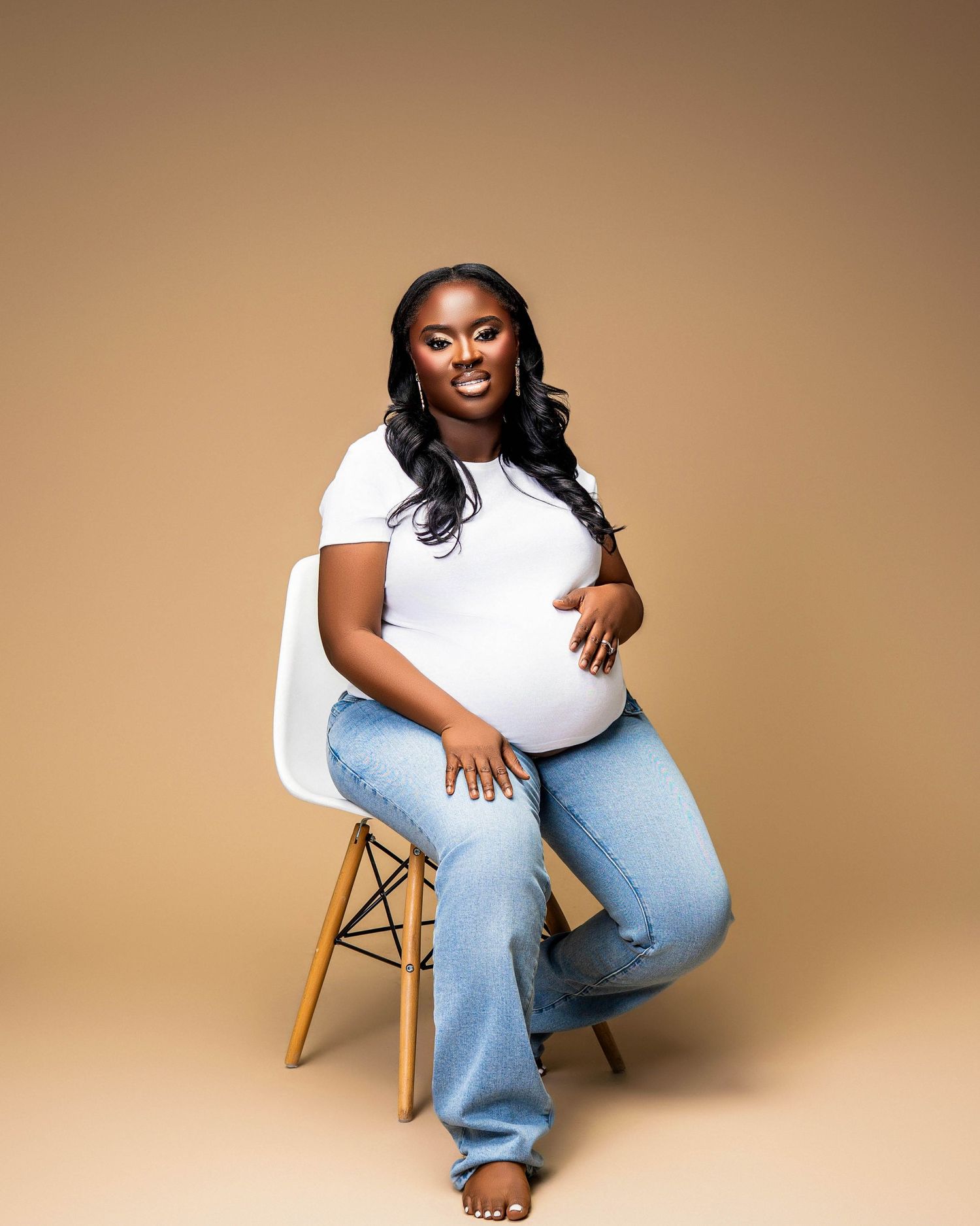 Maternity portrait of woman in white top and jeans seated on wooden stool.