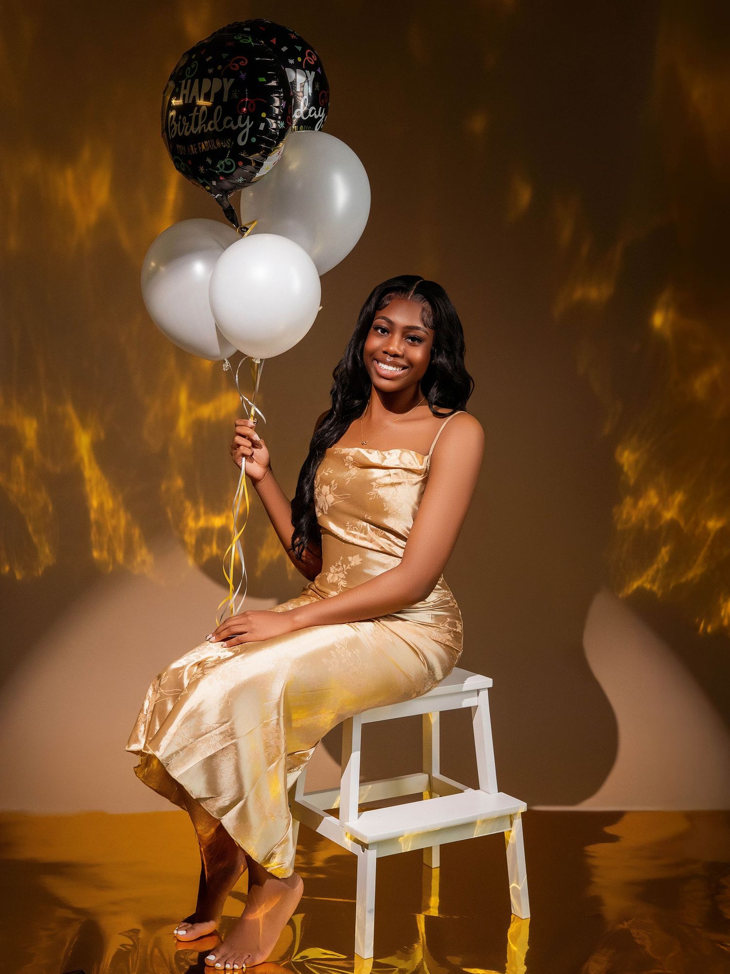 Celebratory portrait in gold satin dress with black and white balloons against warm backdrop.