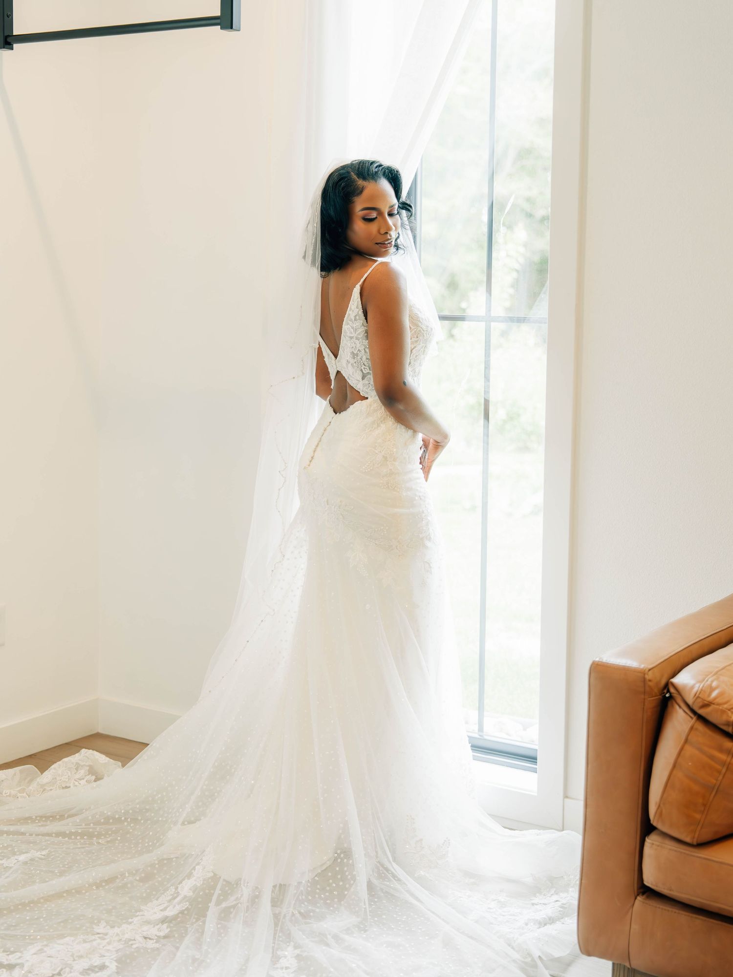 Elegant mermaid wedding dress with lace details and long train shown from behind near window.