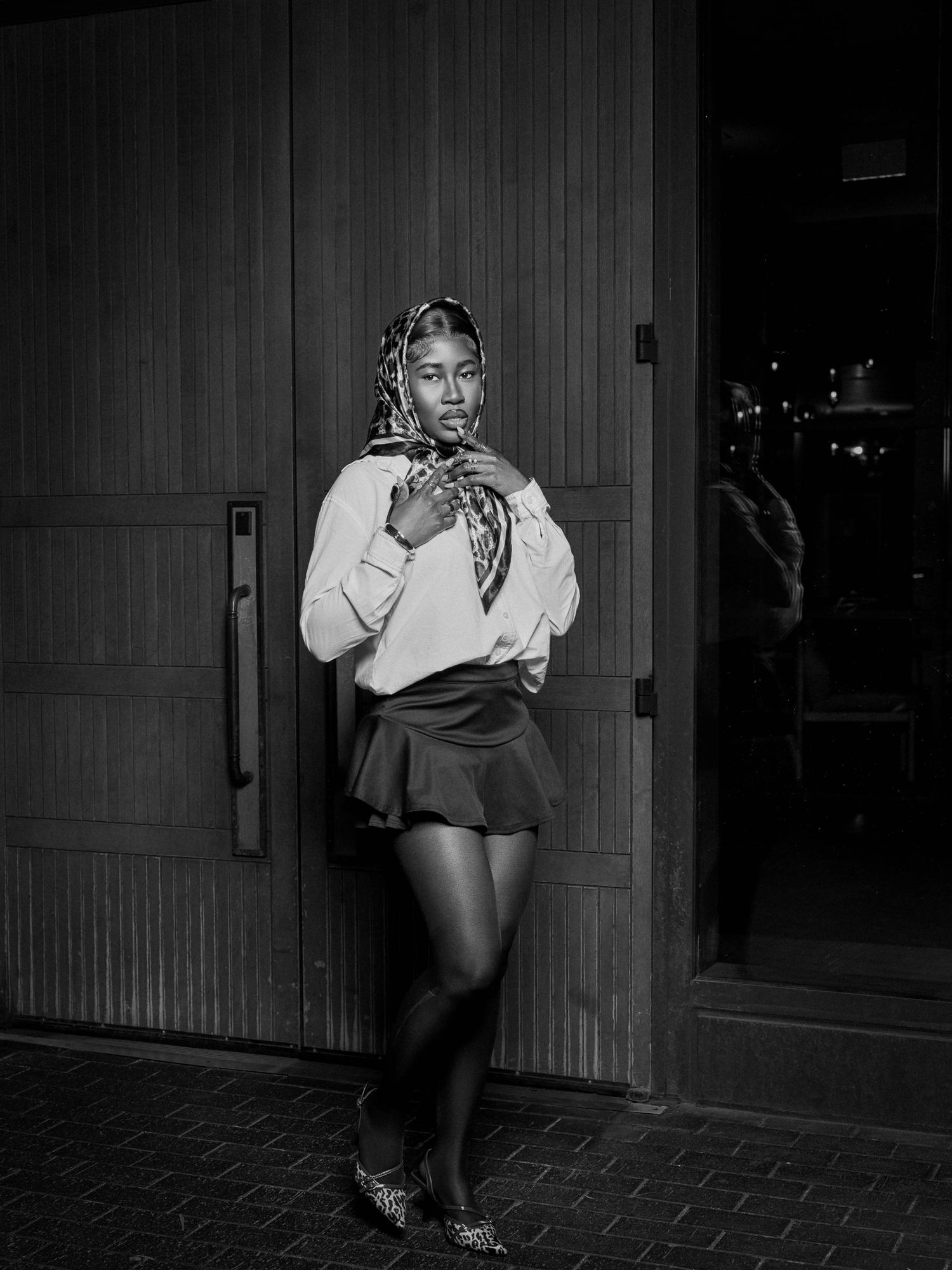 Monochrome fashion photo of a model in a white blouse and patterned headscarf posing outdoors.