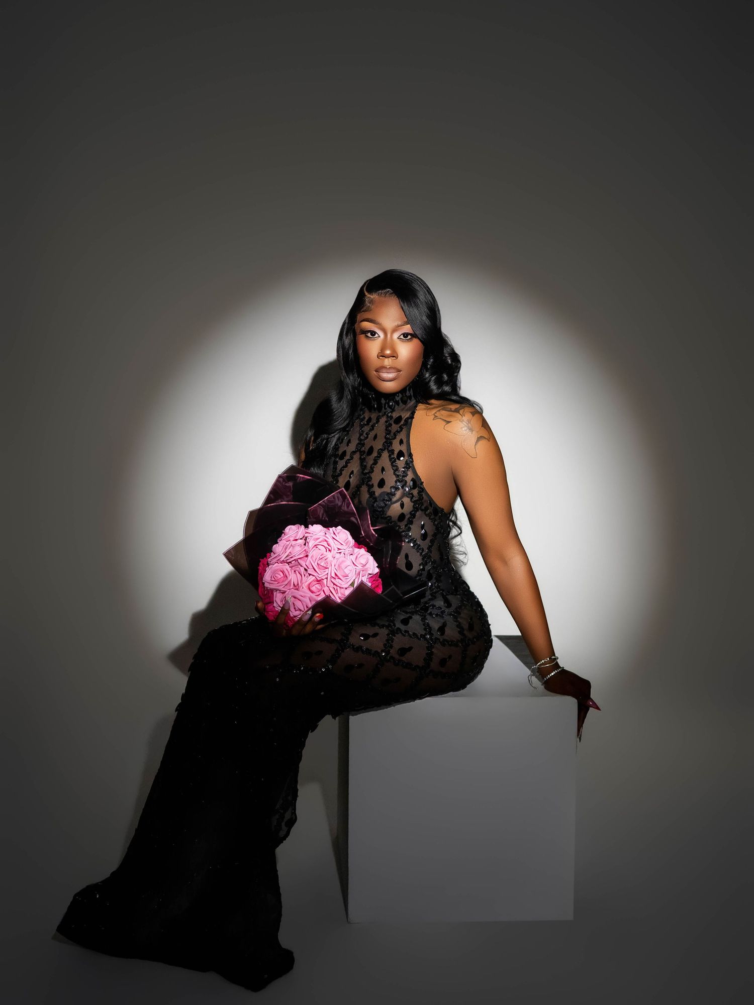 Glamour portrait with black lace gown and pink flowers against dramatic spotlight background.