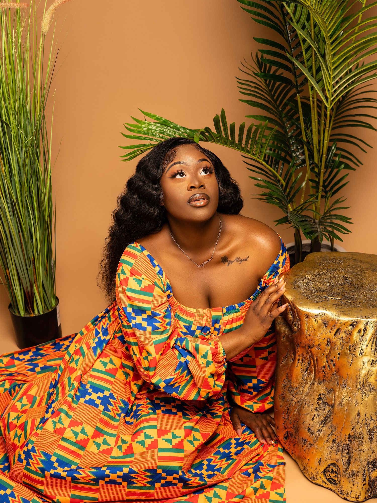A colorful African print dress is styled with tropical palm leaves against a warm brown backdrop in a fashion portrait.