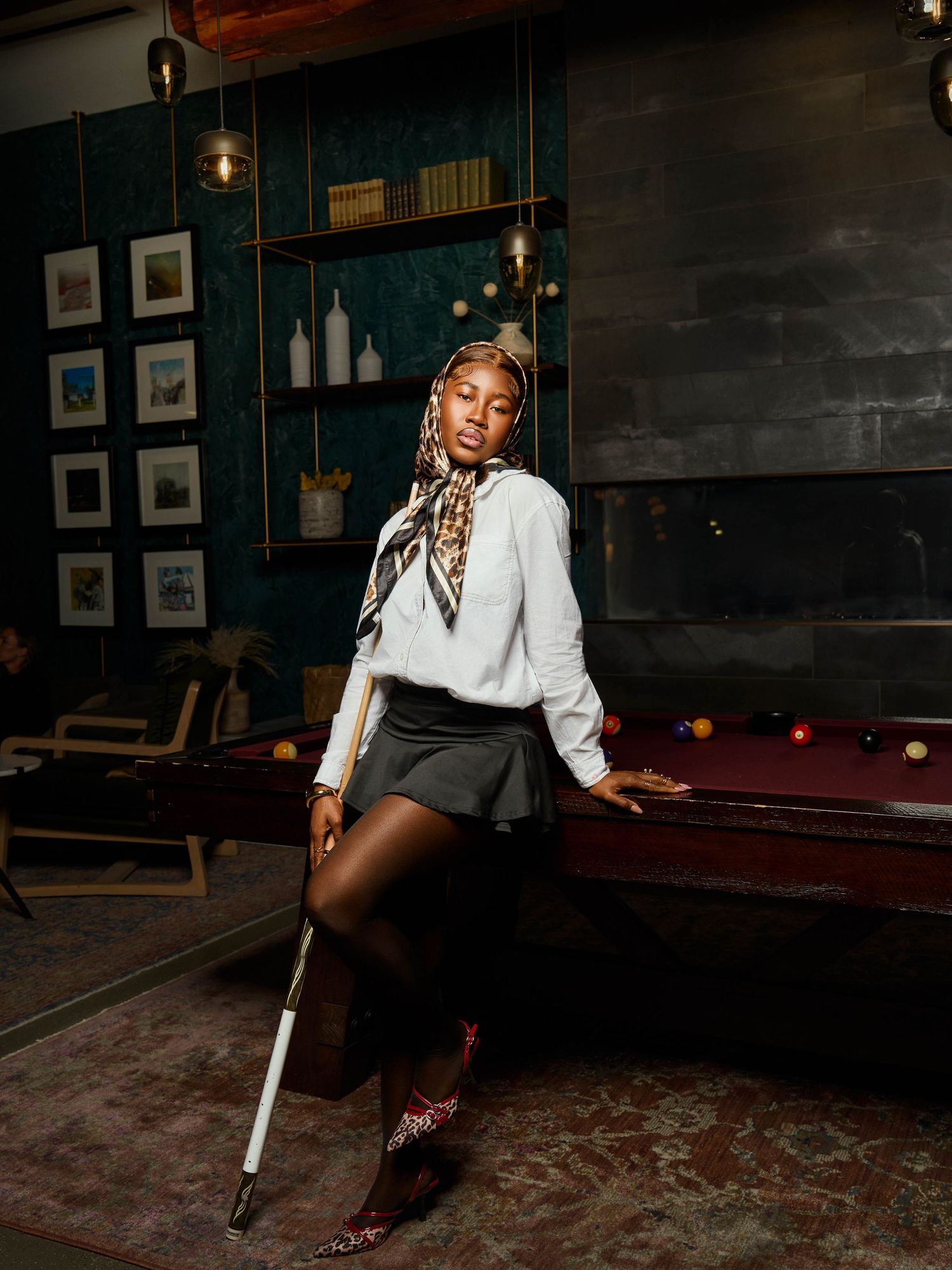 Elegant fashion portrait of a model with a pool cue posing against dark walls and shelving.