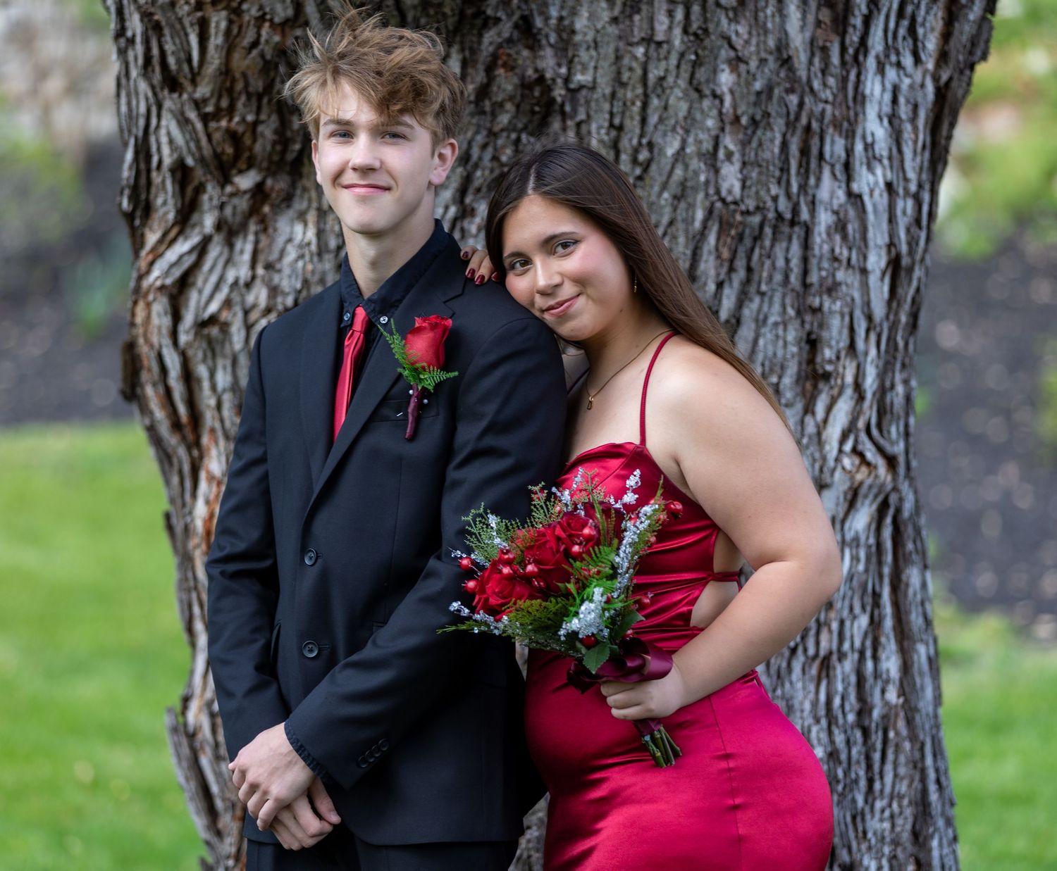 A couple in formal attire poses by a tree trunk - she in a red gown with roses while he wears a dark suit.