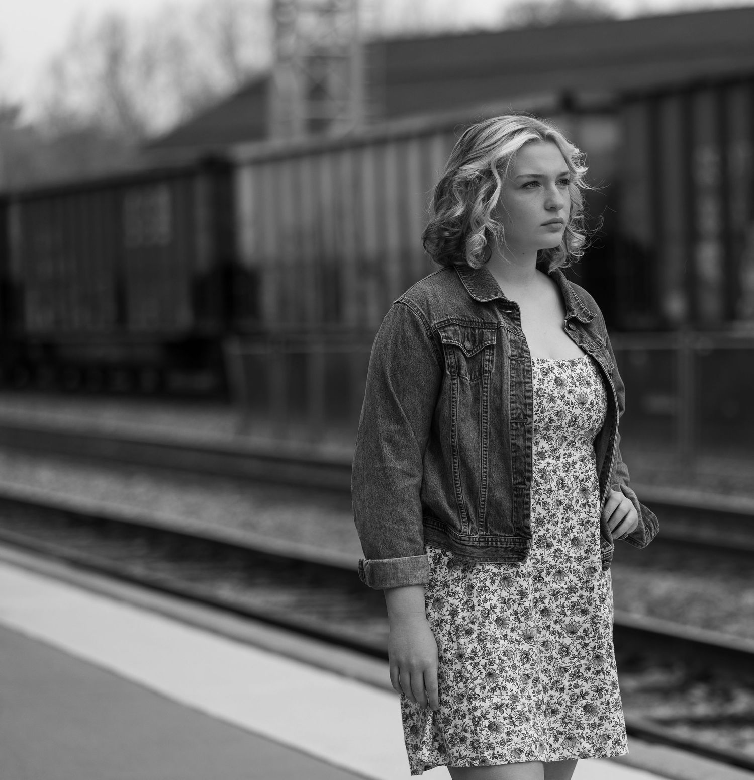 Black and white portrait of a woman in a denim jacket standing by train tracks.