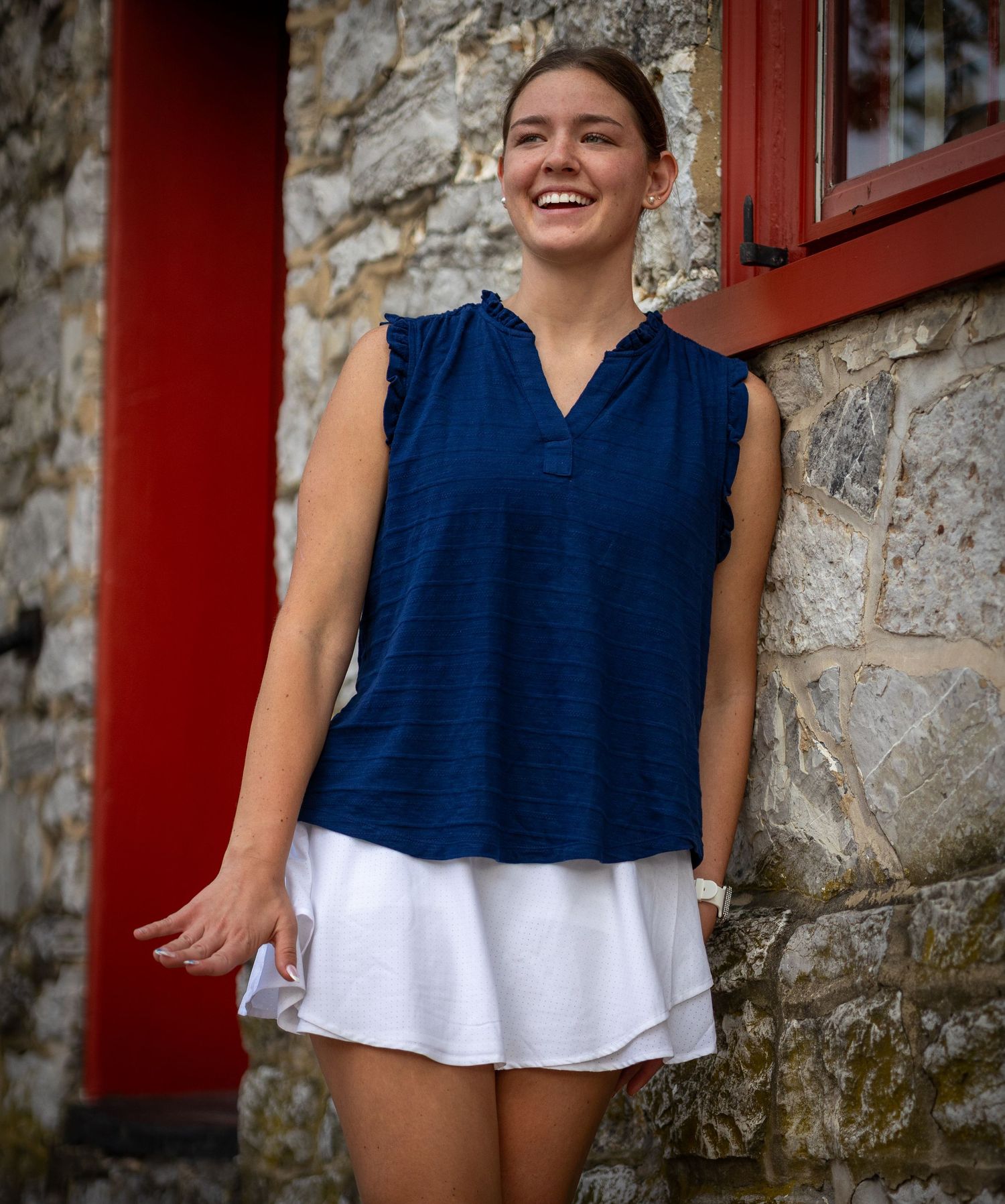Woman in navy sleeveless top and white skirt smiling near red door and stone wall.