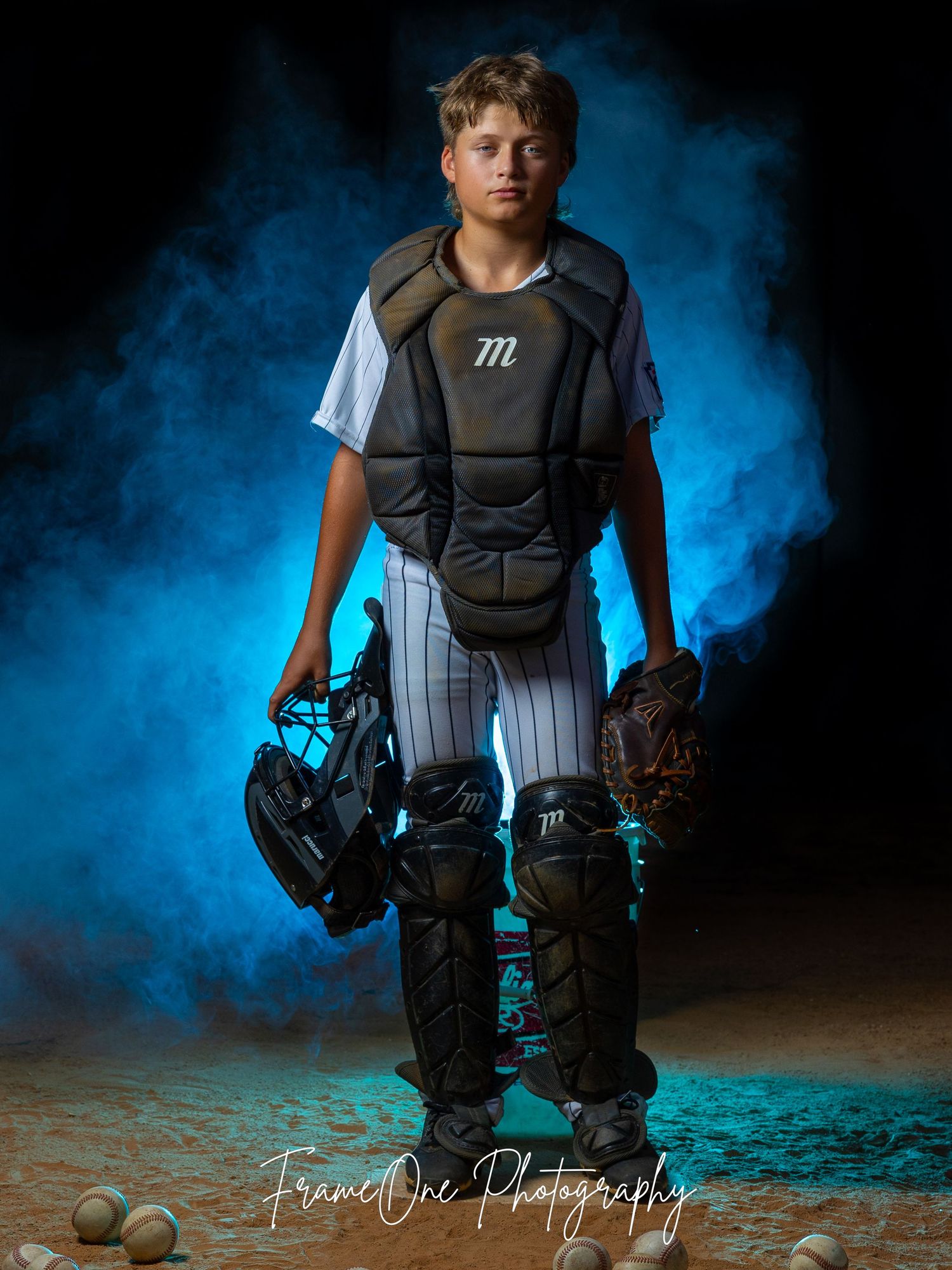 Youth baseball catcher in protective gear poses against dramatic blue smoke background during sports photoshoot.