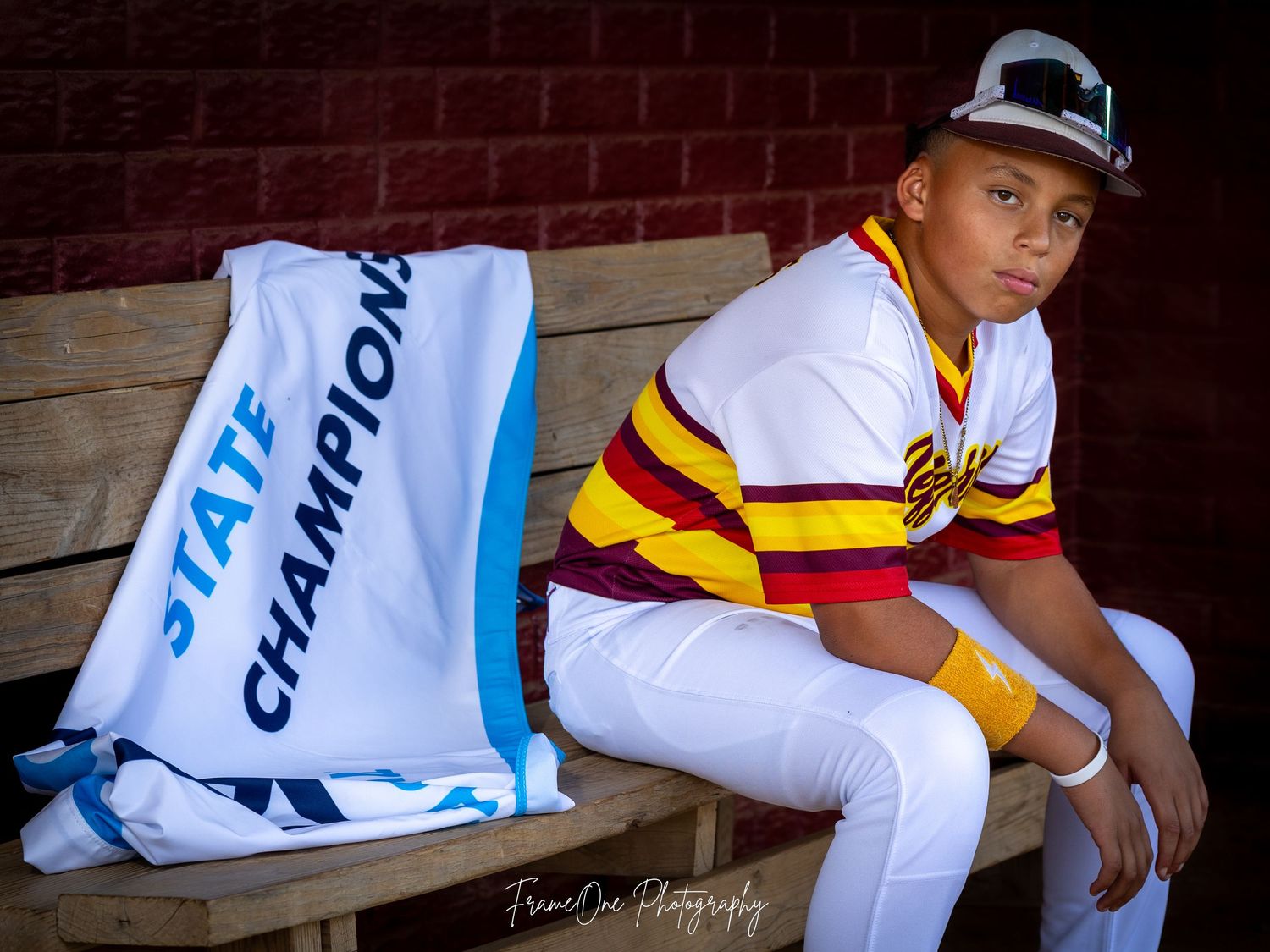 Baseball player in white and yellow striped uniform sits on dugout bench next to light blue championship towel.