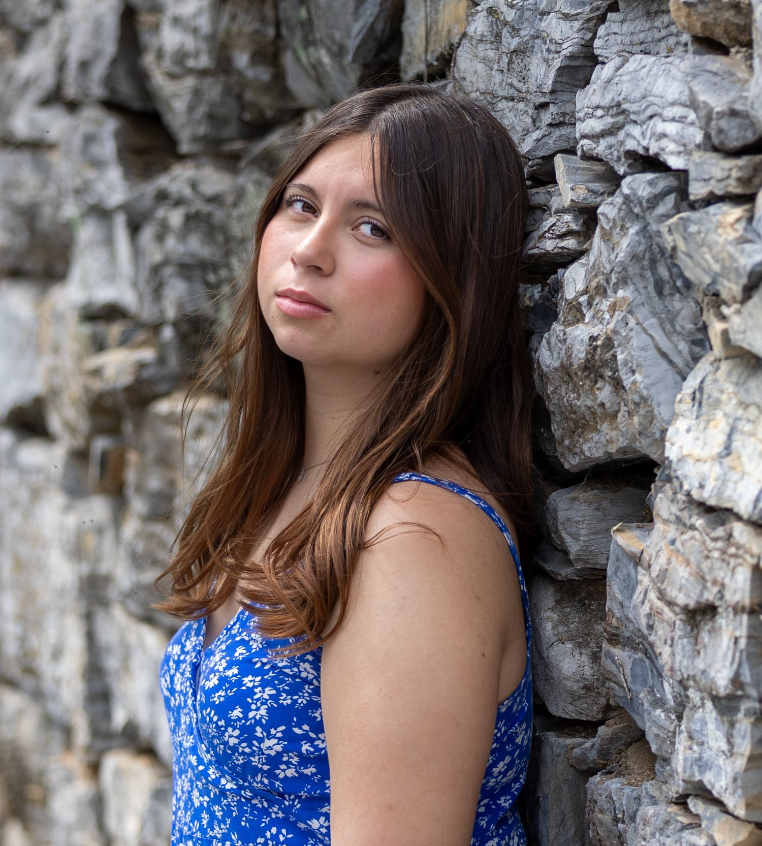 Young woman in blue floral dress poses against textured stone wall background for outdoor portrait session.