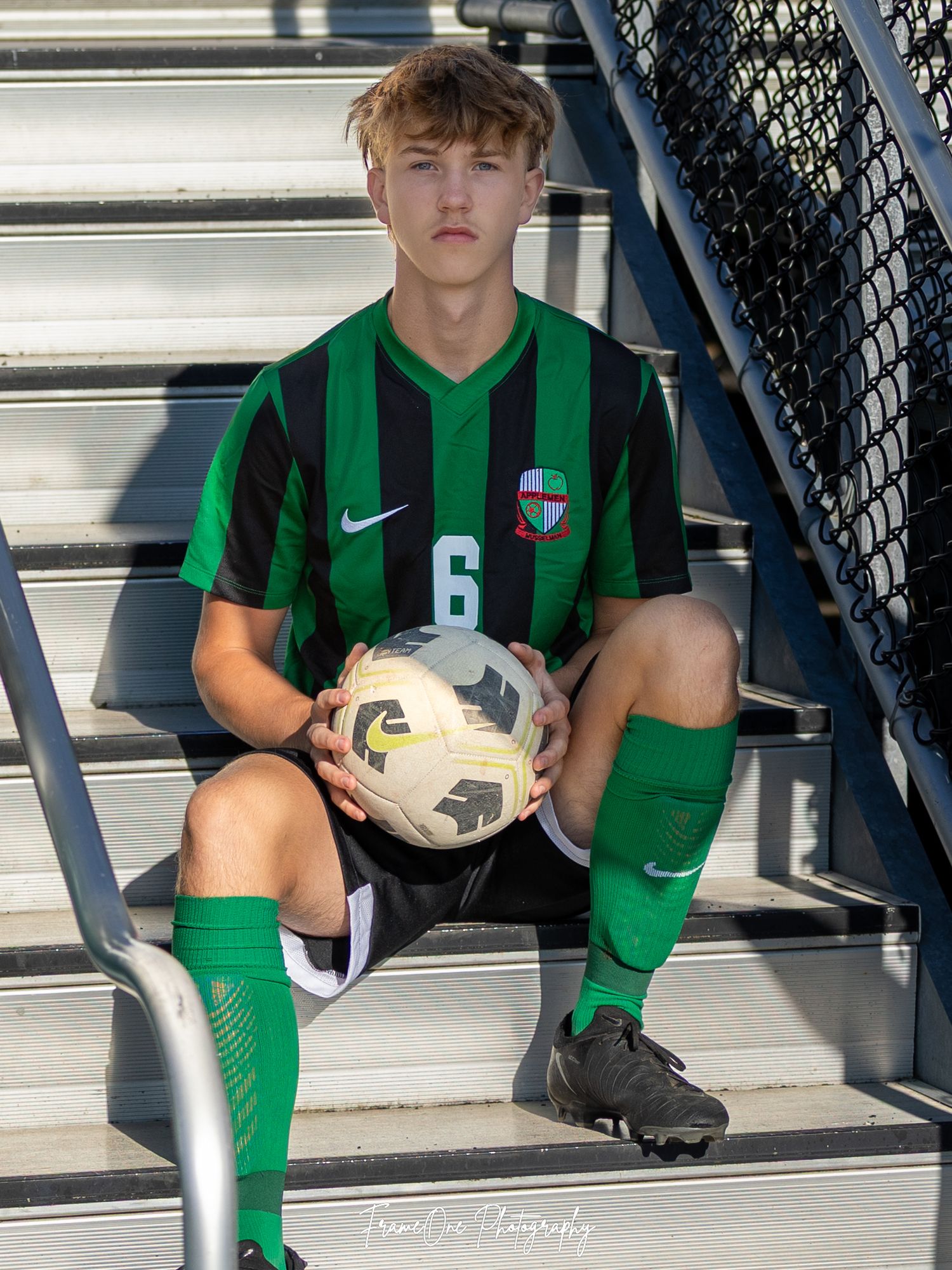 Soccer player in green and black striped uniform sits on stadium bleachers holding a ball.