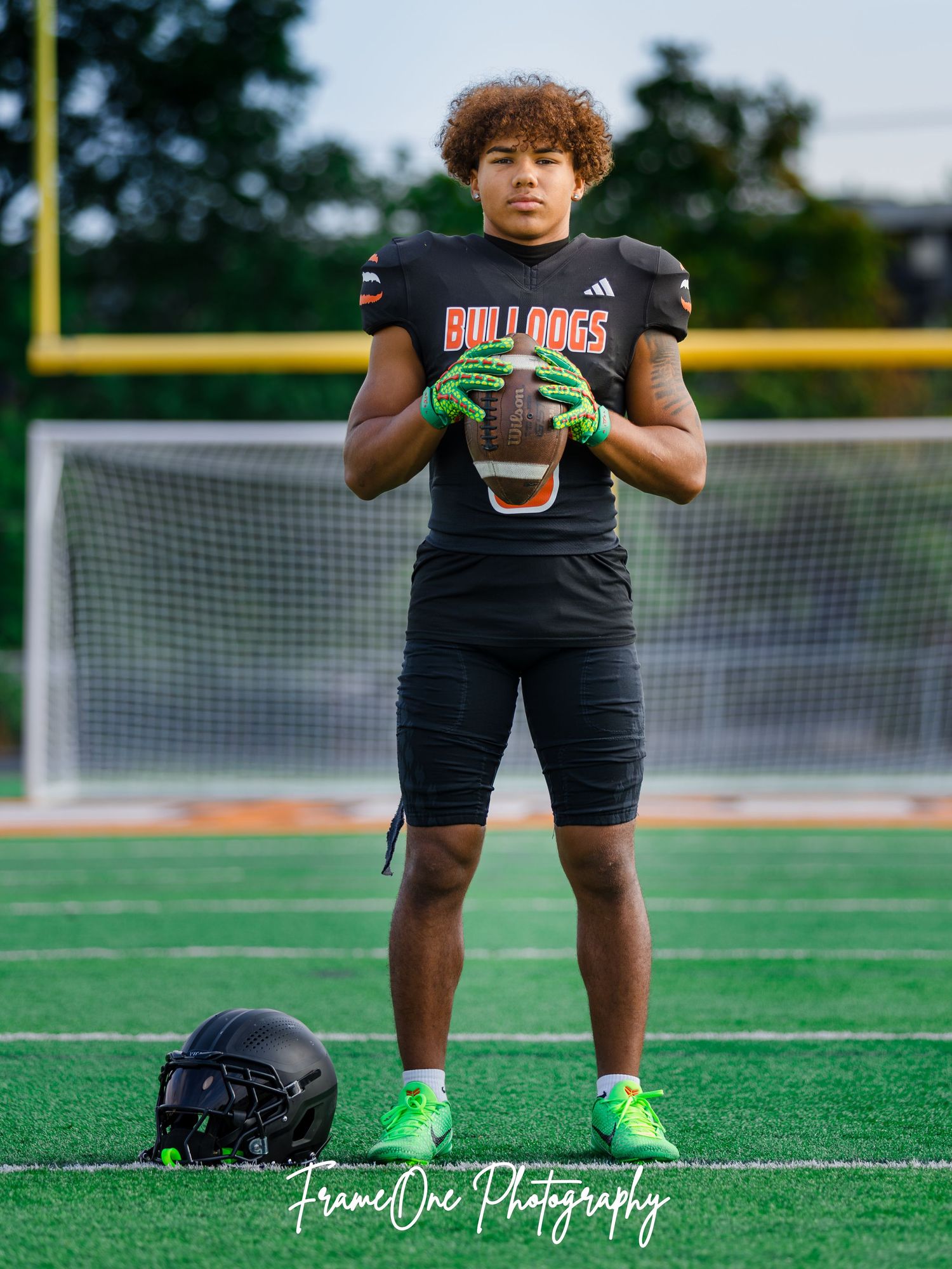 High school football player in black uniform stands on field with bright green cleats and gloves while preparing for practice.