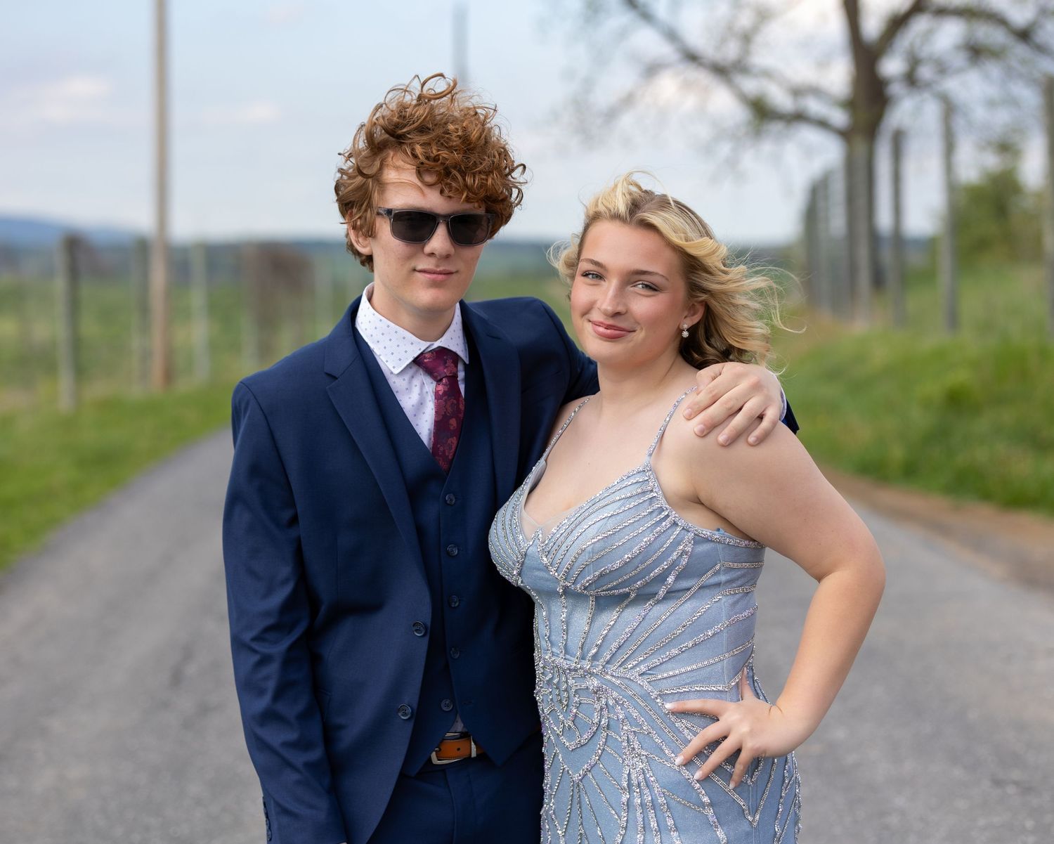 A formally dressed couple poses together on a rural road with trees and countryside in the background.