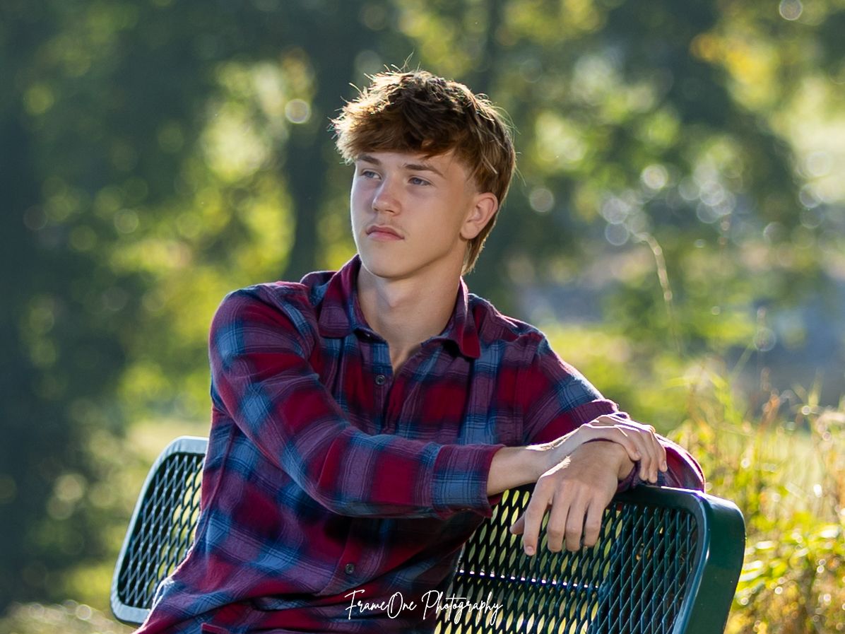 Outdoor senior portrait of student in red and blue plaid shirt sitting on bench with sunlit nature background.