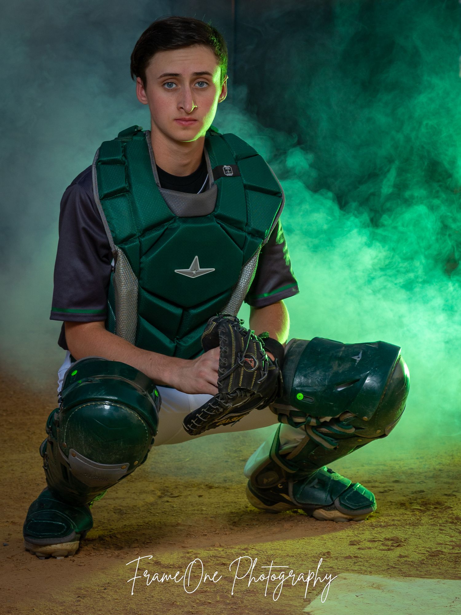Baseball catcher crouches in protective gear against dramatic green smoke background during studio photoshoot.