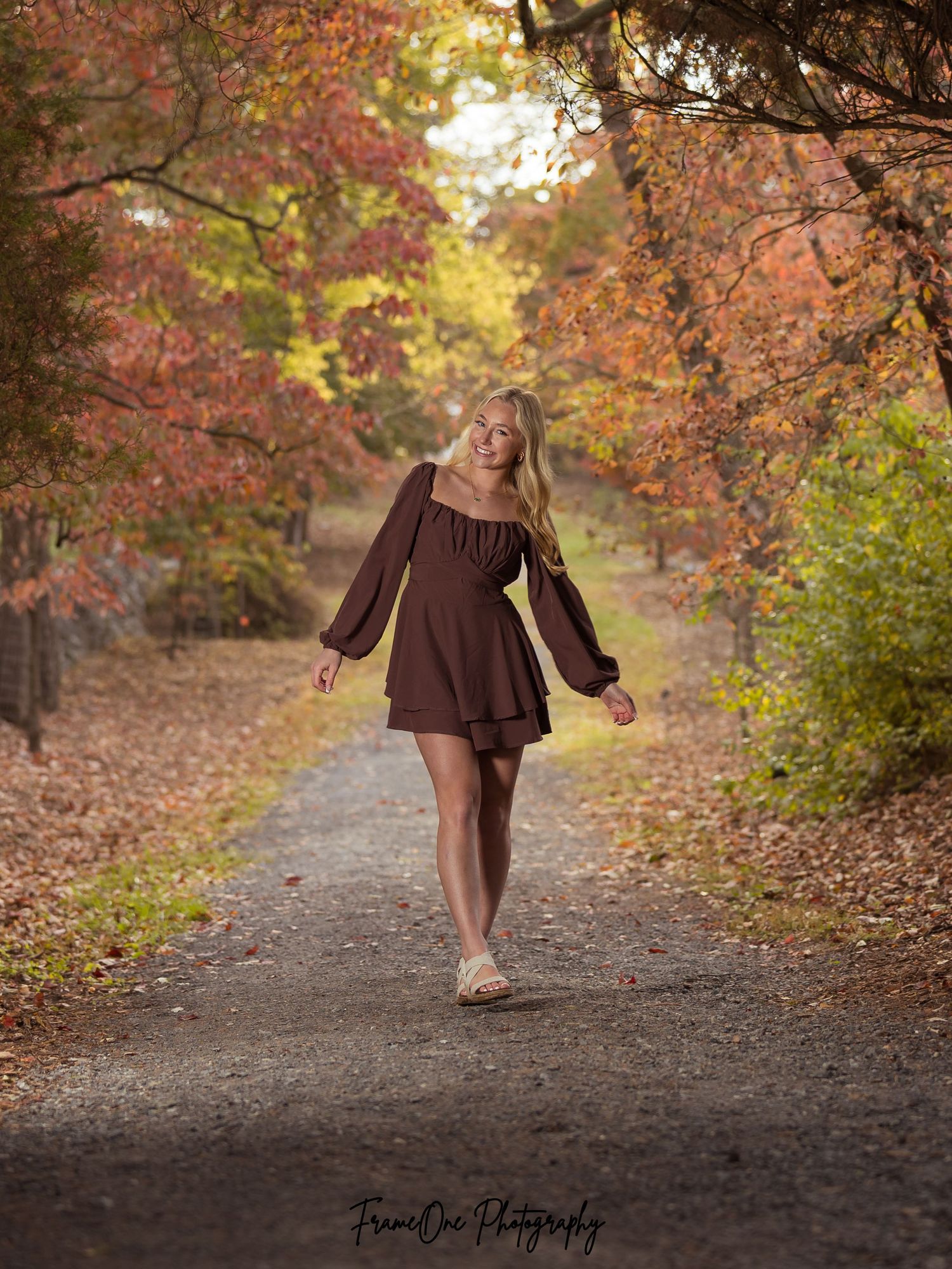 A person in a brown dress twirls on a dirt path surrounded by autumn trees with golden and red foliage.