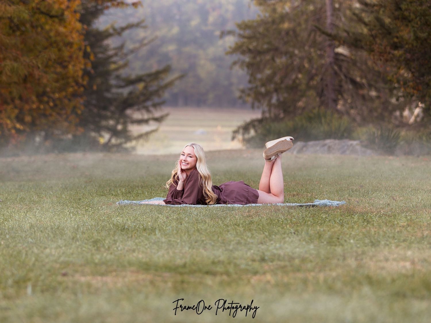 Someone lying on a blanket in a misty field at sunset with pine trees in the background.