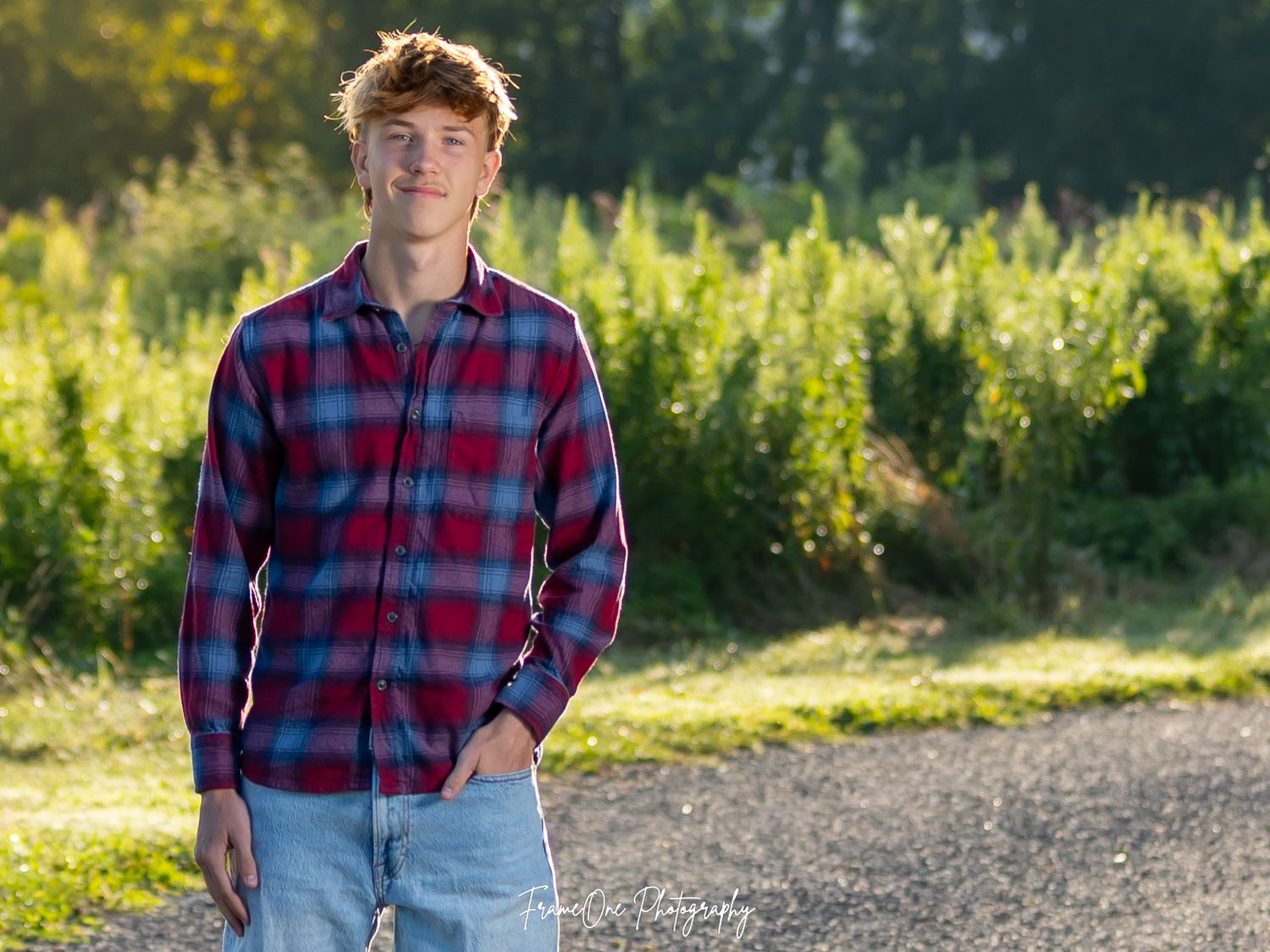 A person wearing a red and navy plaid shirt poses outdoors in a sunny field with golden wildflowers in the background.